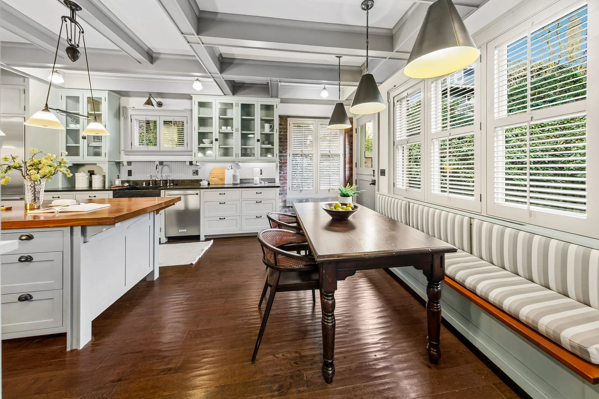 Bright kitchen and dining area with large windows, white cabinetry, wooden table, striped built-in bench, and hanging pendant lights.