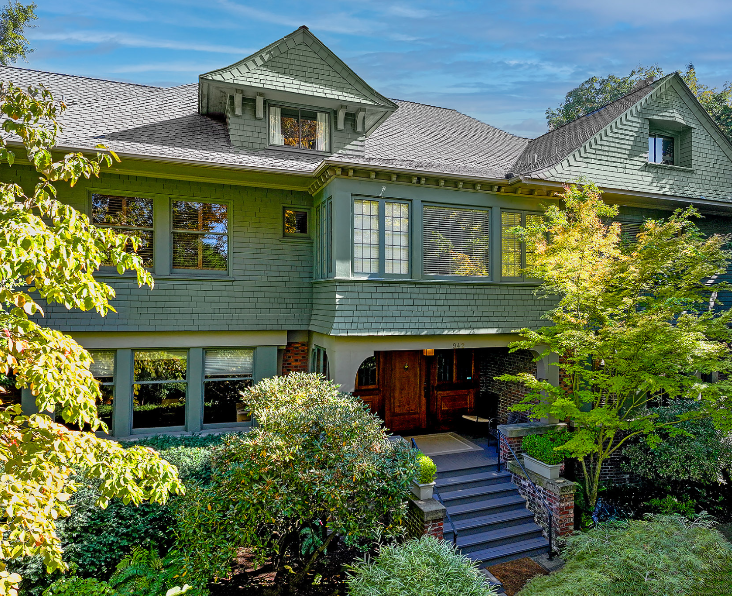 Front view of a large, green, three-story house with a steep roof, multiple windows, and a wooden front door, surrounded by lush greenery and trees under a blue sky.