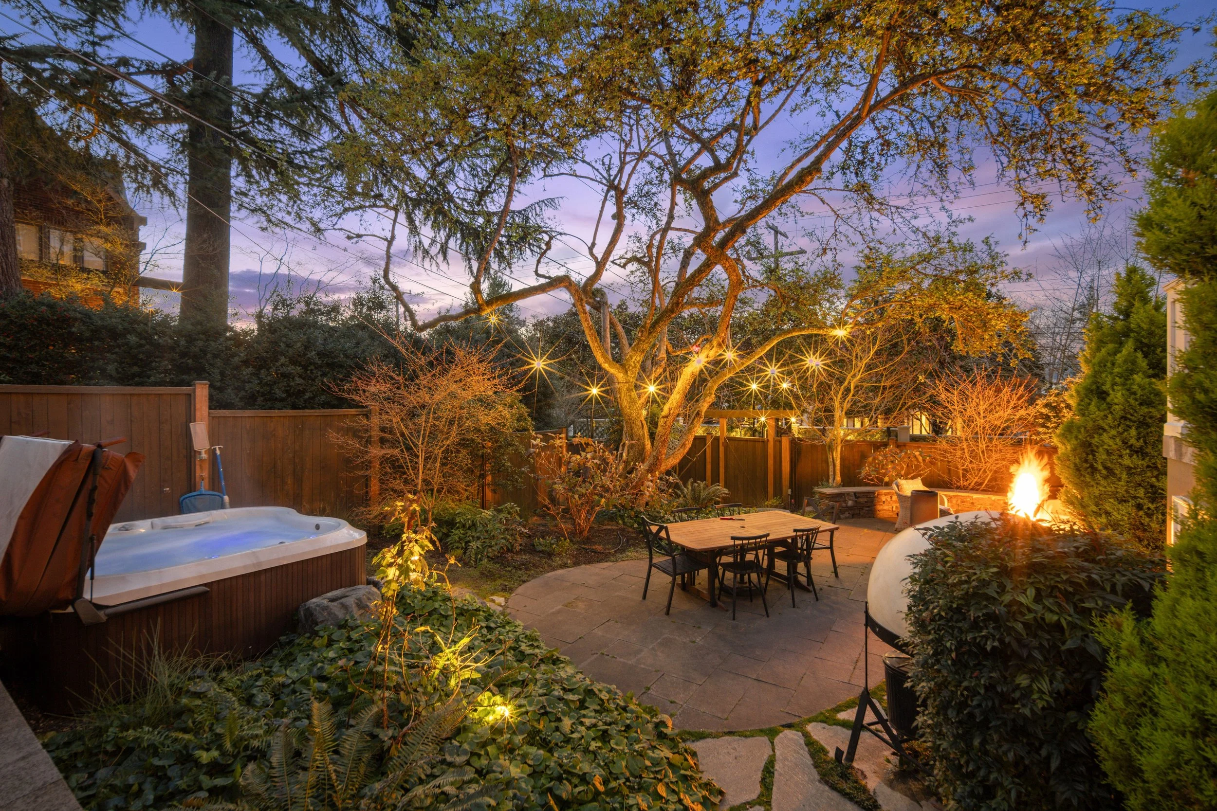 A backyard patio with string lights hanging from trees, a hot tub on the left, and a fire burning on the right, surrounded by trees and a wooden fence.