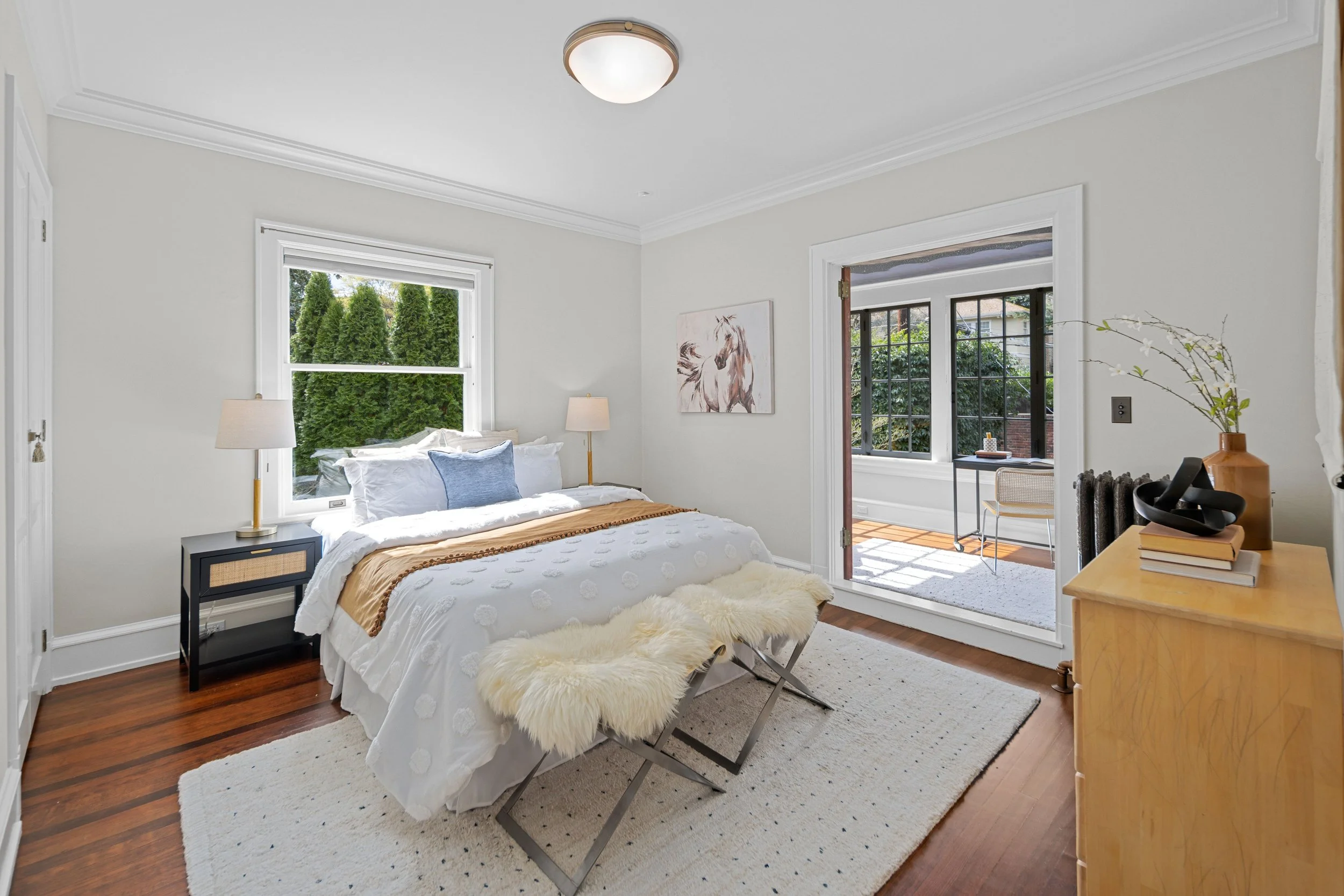 Bright bedroom with white walls, bed with white bedding and blue pillows, two bedside tables with lamps, hardwood floors, and a view of a sunlit enclosed porch with large windows.