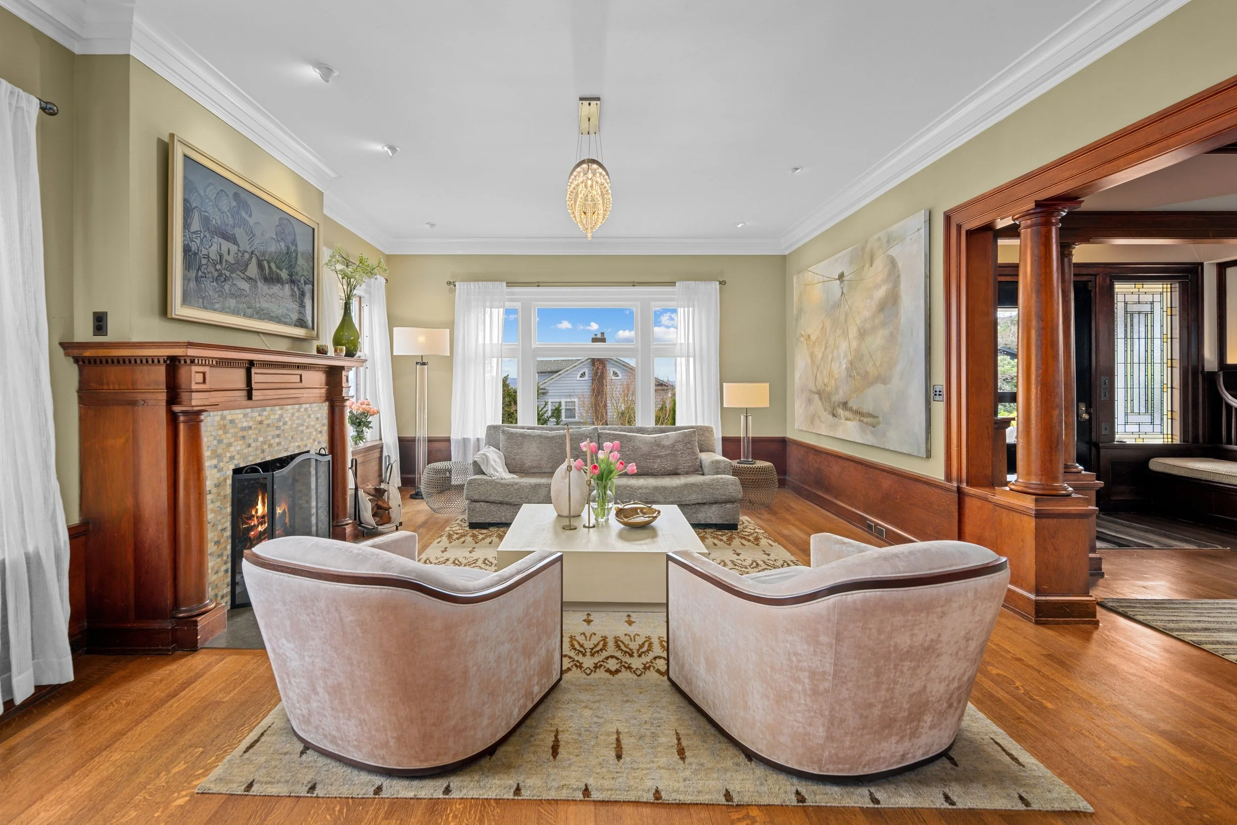 Living room with a fireplace, two pink armchairs, a gray sofa, and large window with white curtains, decorated with artwork, lamps, and a coffee table with pink tulips.