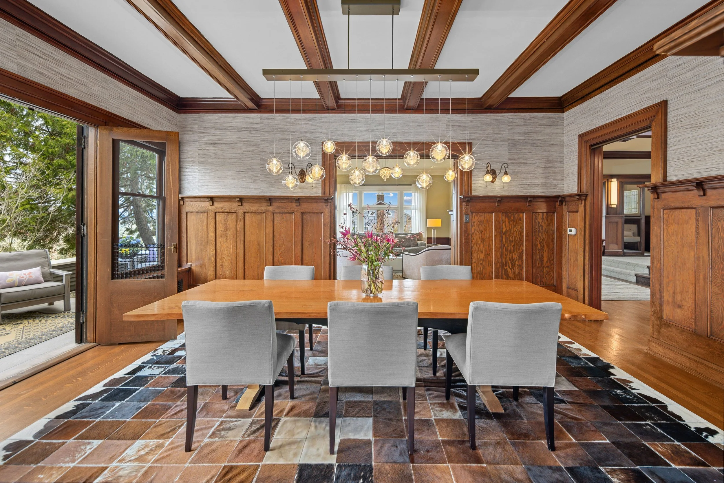 A wooden dining table with six light gray chairs in a room with wood-paneled walls and a large window. There is a modern hanging light fixture and a vase with pink flowers in the center of the table. The room opens to an outdoor patio with a bench an