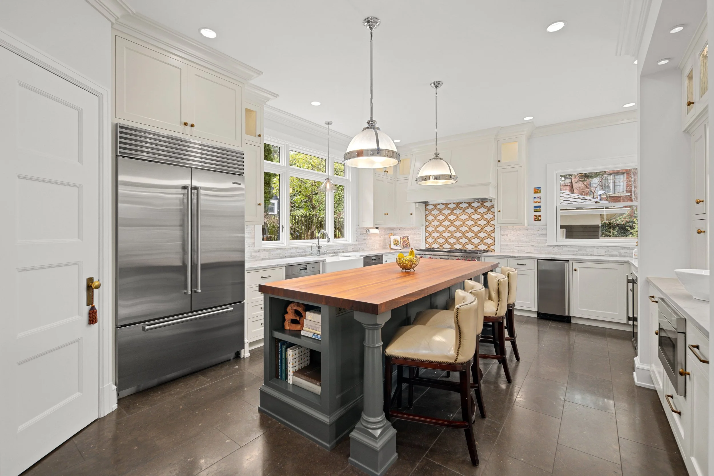 A spacious modern kitchen with white cabinets, stainless steel appliances, a large wooden island with cream-colored chairs, large windows, and a patterned tile backsplash.