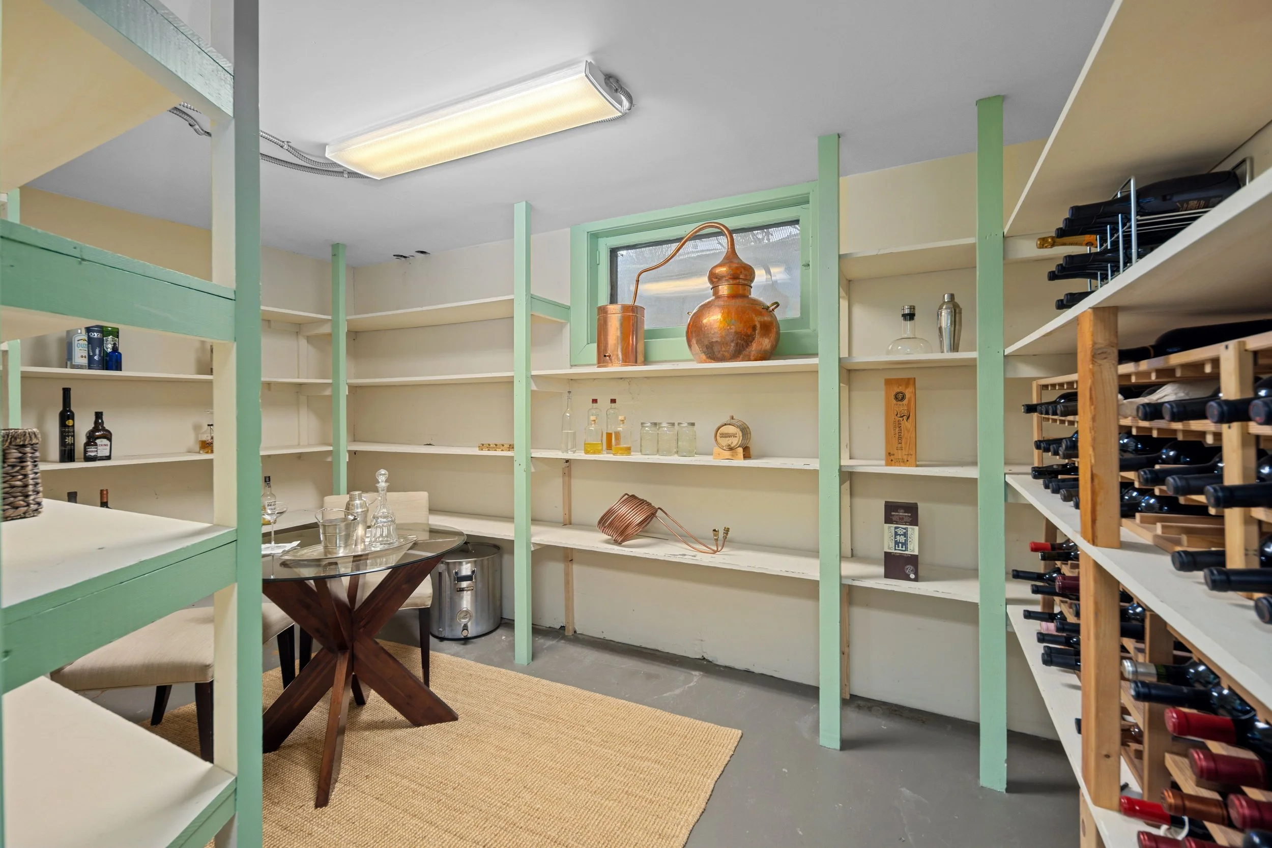 A walk-in wine cellar with multiple shelves holding bottles and decanters, a small table with glassware, a beige rug, and a small window with copper distillation apparatus on the windowsill.