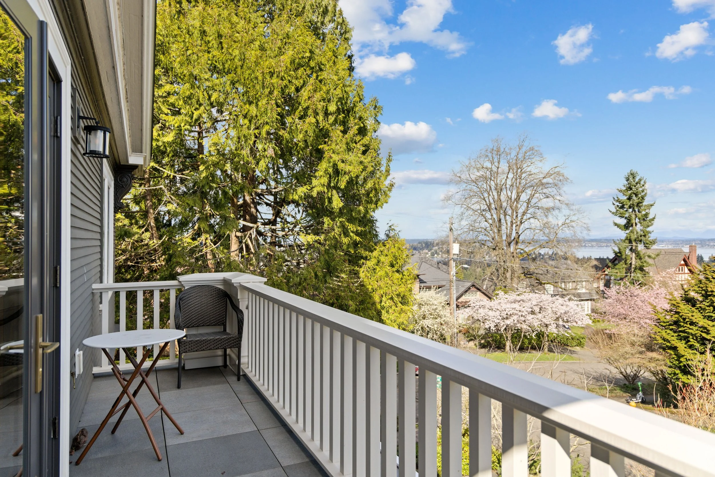 A balcony with a small round white table and a black wicker chair, overlooking a suburban neighborhood with trees, houses, and a body of water in the distance under a partly cloudy sky.