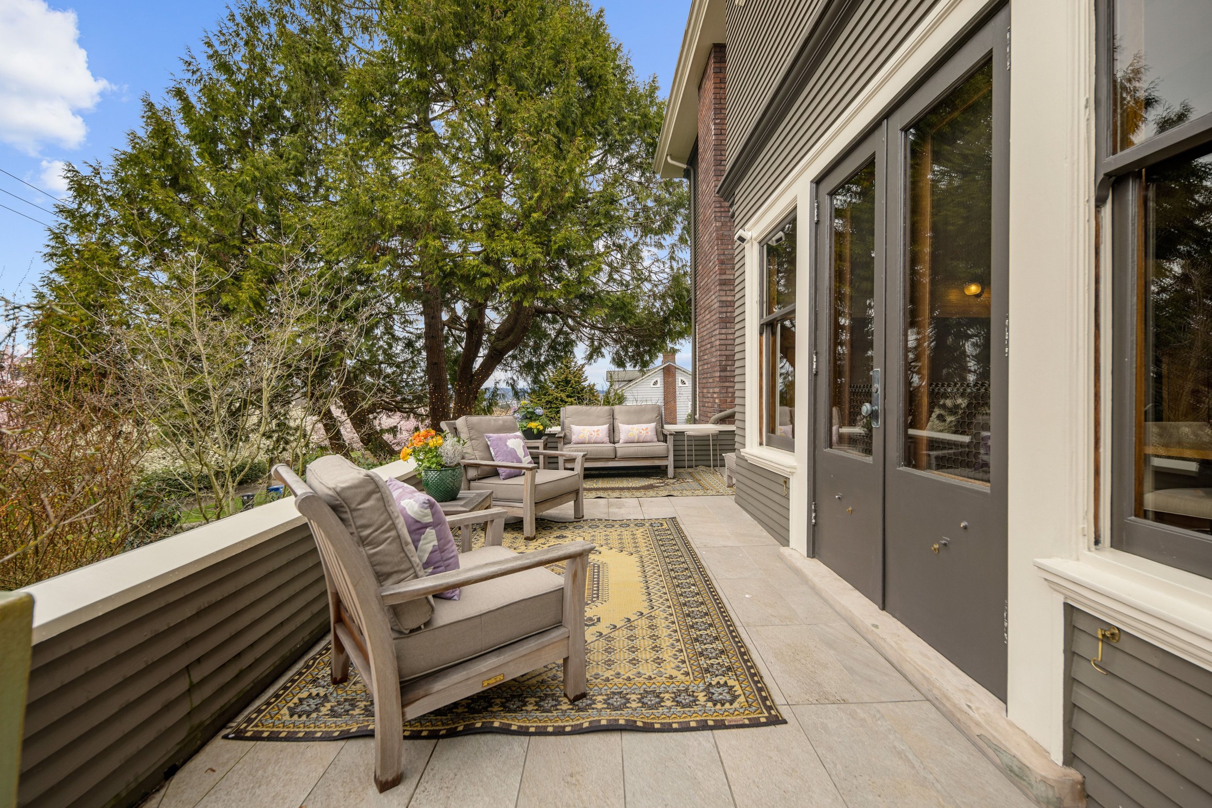Outdoor patio with chairs, potted plants, colorful cushions, and a large tree in the background.