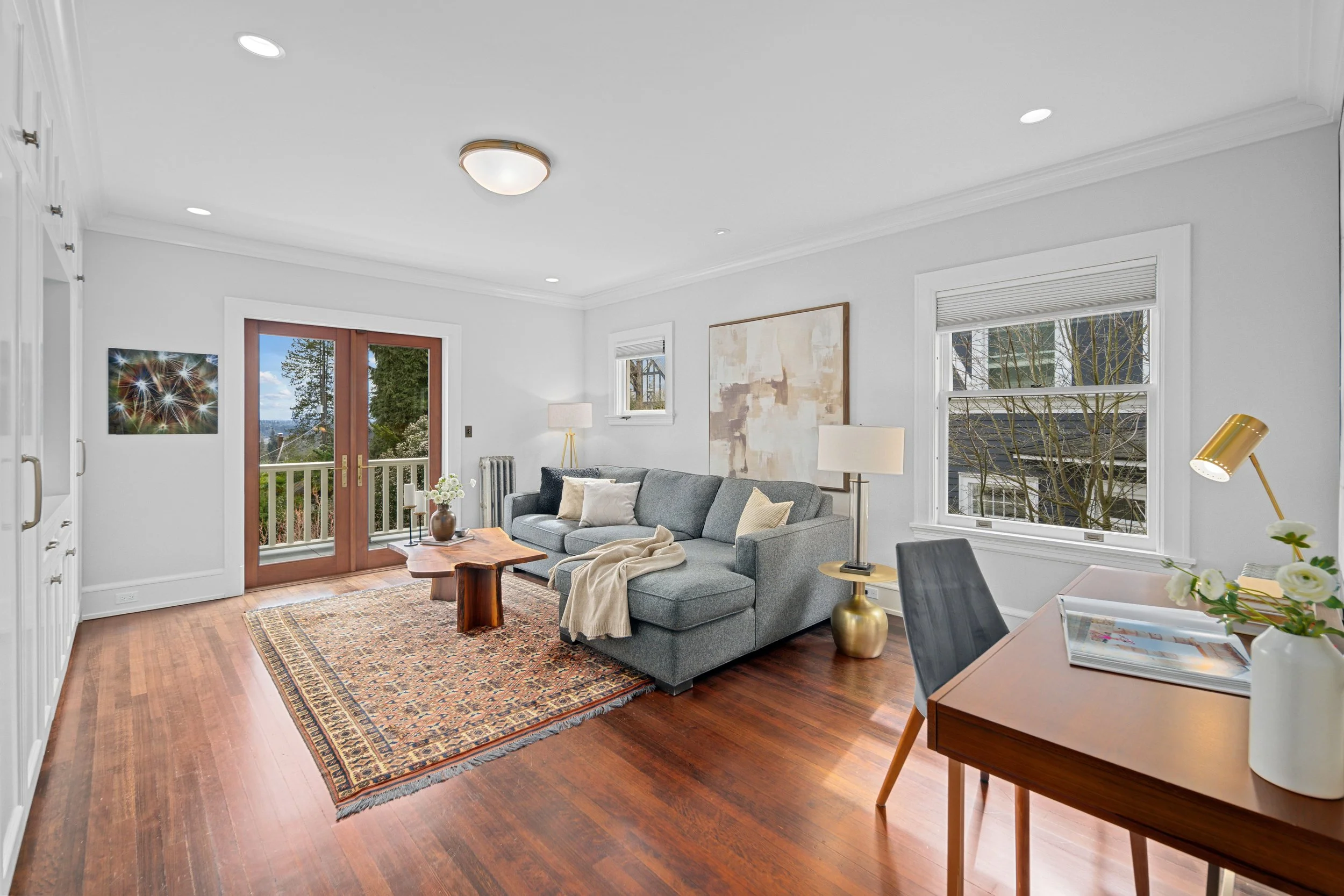 Bright living room with grey sofa, wooden coffee table, patterned area rug, large windows with blinds, and door leading to a balcony with trees outside.