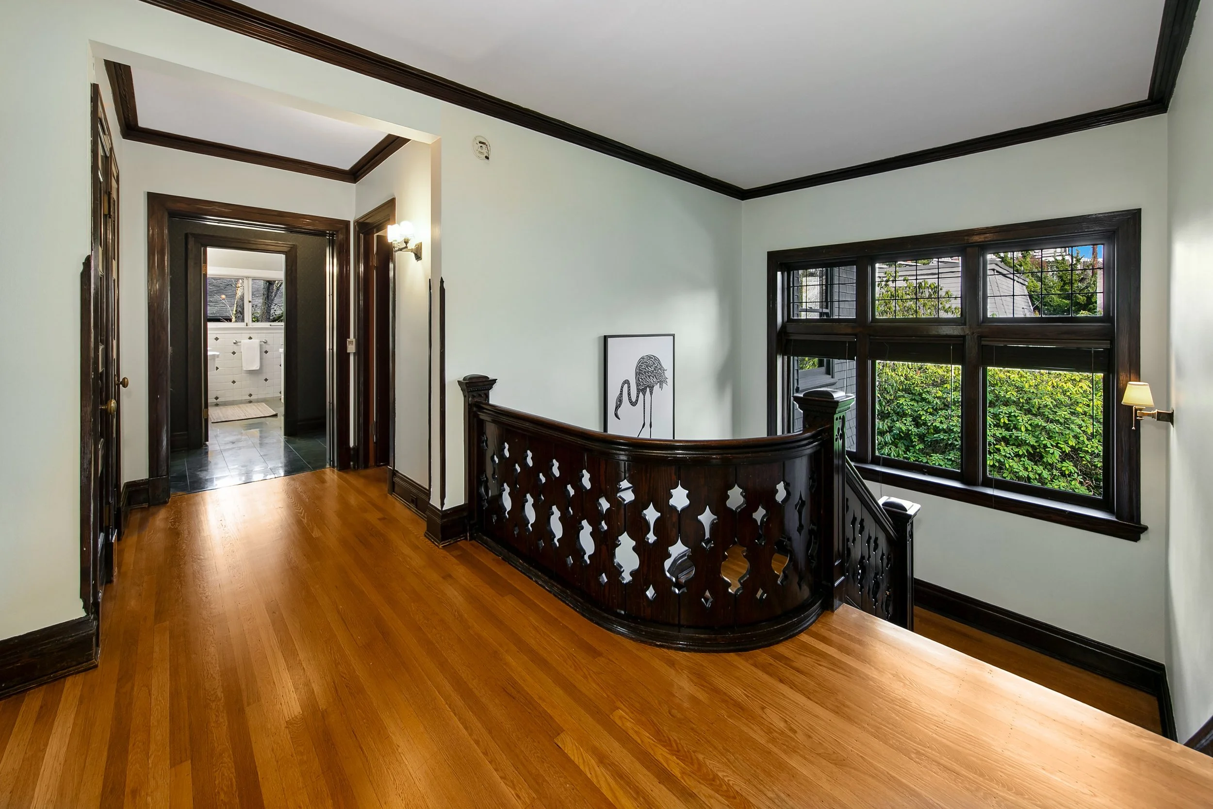 Hallway with wooden floors and dark wood trim, leading to a bathroom. A staircase with dark wood banister and decorative panels curves along the wall, and windows with dark wood framing look outside to greenery.