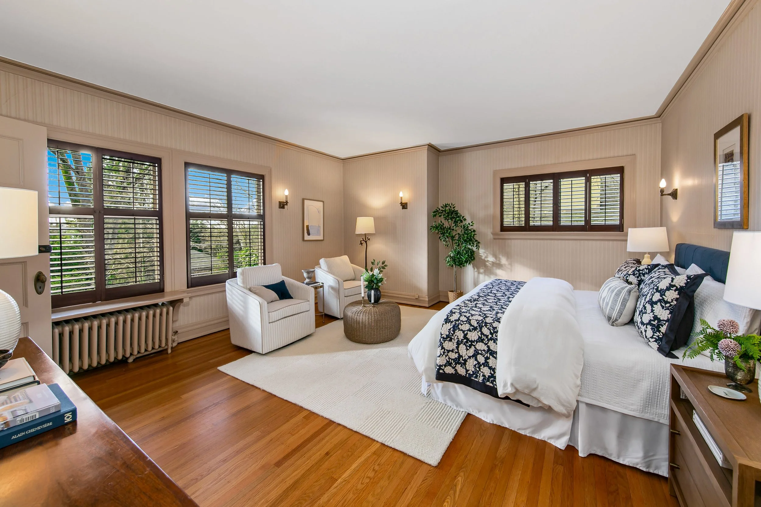 A bright bedroom with beige walls, hardwood floors, and multiple windows with wooden blinds. It features a white bed with patterned pillows, a small sitting area with armchairs, a round ottoman, and various lamps and decorations.