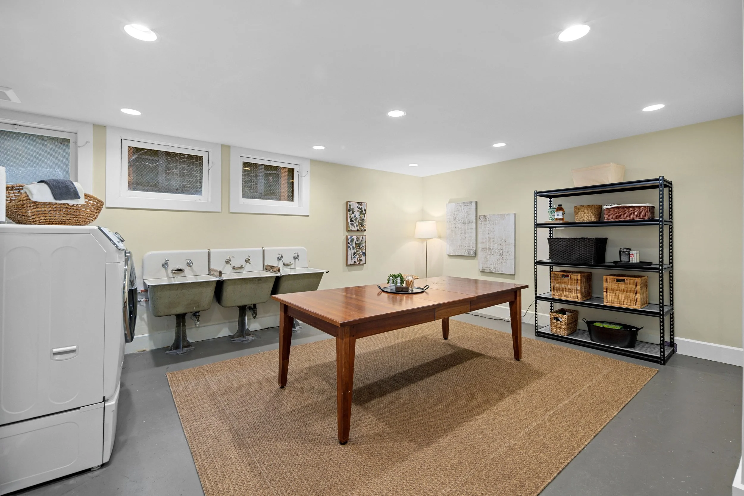 Laundry room with washing machine, double utility sinks, wooden table, black metal shelf with baskets, and a wall lamp.