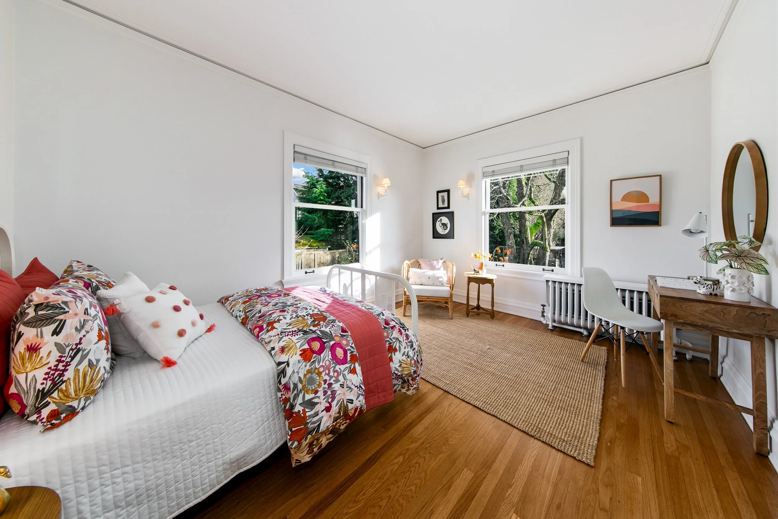 A bright, cheerful bedroom featuring a bed with colorful floral and pink bedding, two large windows with a view of greenery, a small sitting area with a chair and side table, a wooden desk with a white chair, and walls decorated with pictures and a m