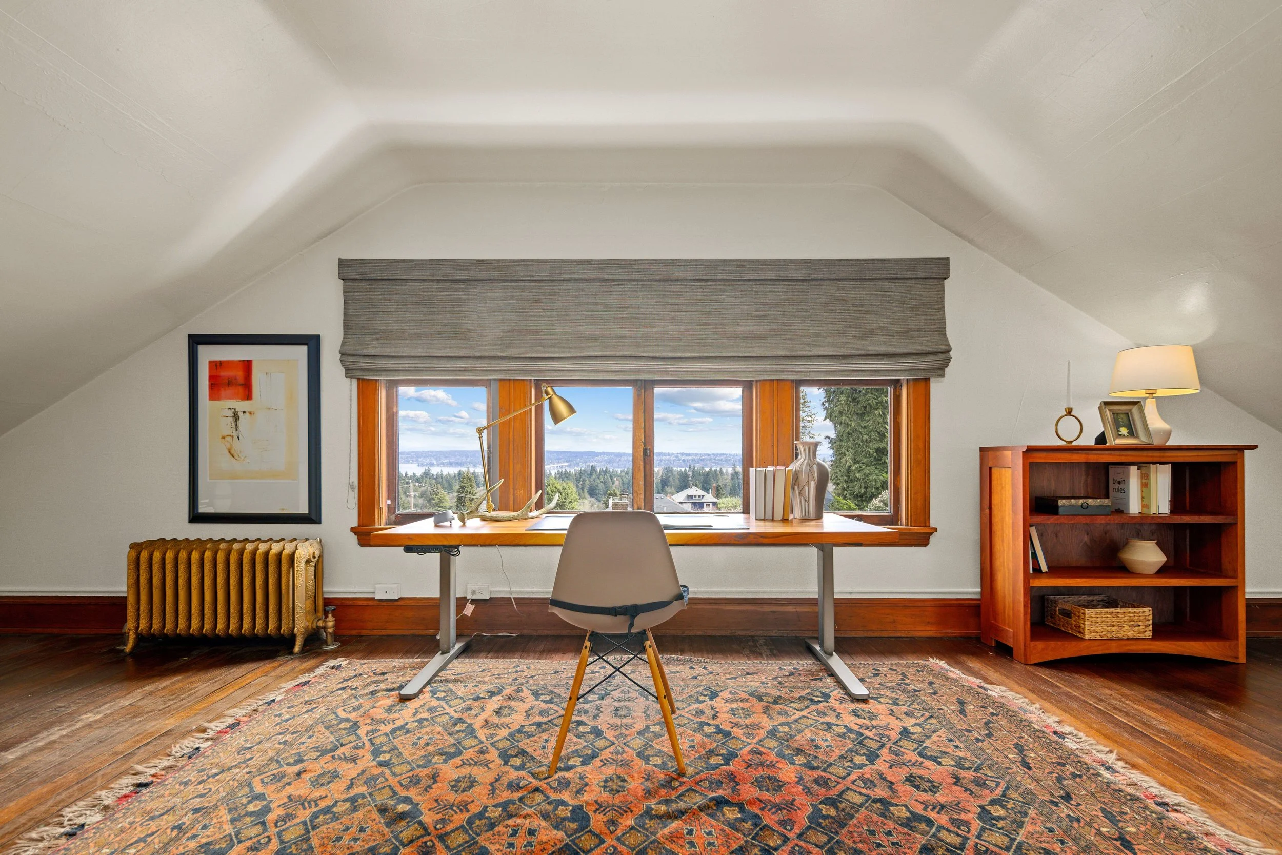 Home office with desk facing large window, beige chair, bookshelf, and decorative objects, with spacious wooden floor and mountain view.