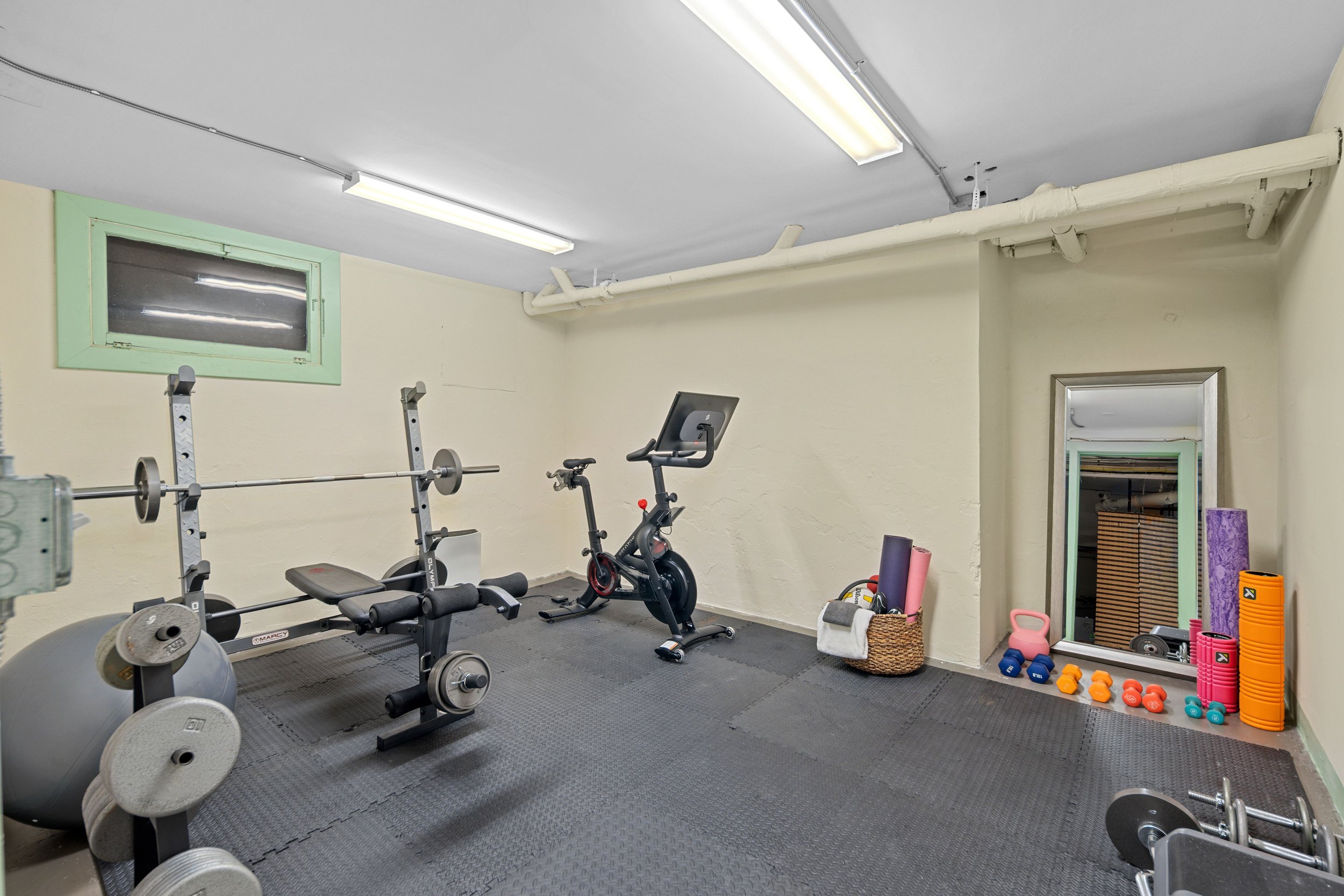 Home gym with workout equipment including a weight bench, barbell, stationary bike, dumbbells, foam rollers, yoga mats, weight plates, and a mirror on the wall.