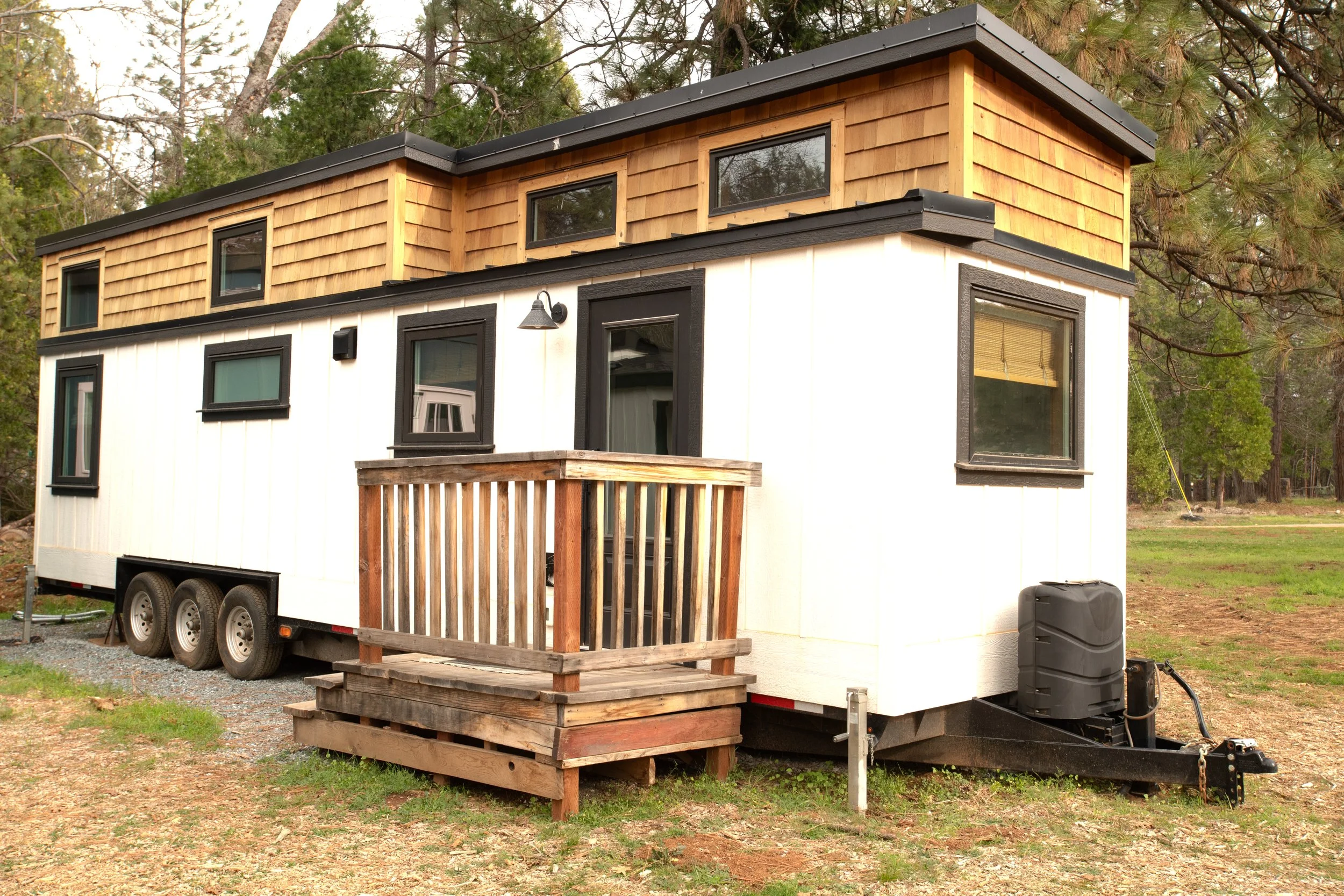 A modern tiny house on wheels with white and wooden exterior, small front porch, black framed windows, surrounded by trees and grass.
