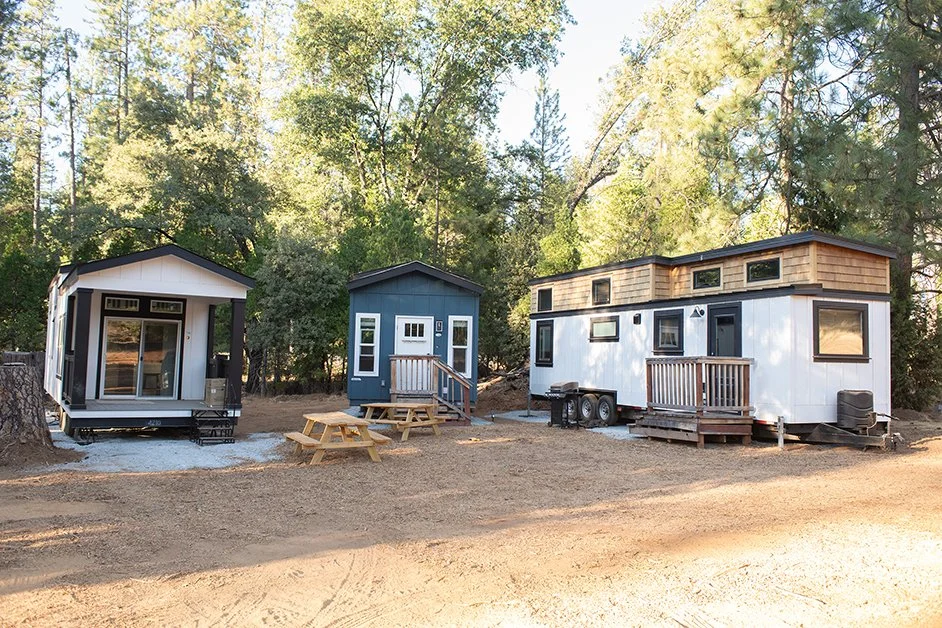 Three tiny houses with different designs and colors are situated in a wooded area, with trees in the background. The house on the left is black and white, the middle house is blue, and the right house is white with wooden accents. There are small wooden decks in front of each house, with picnic tables placed outside.