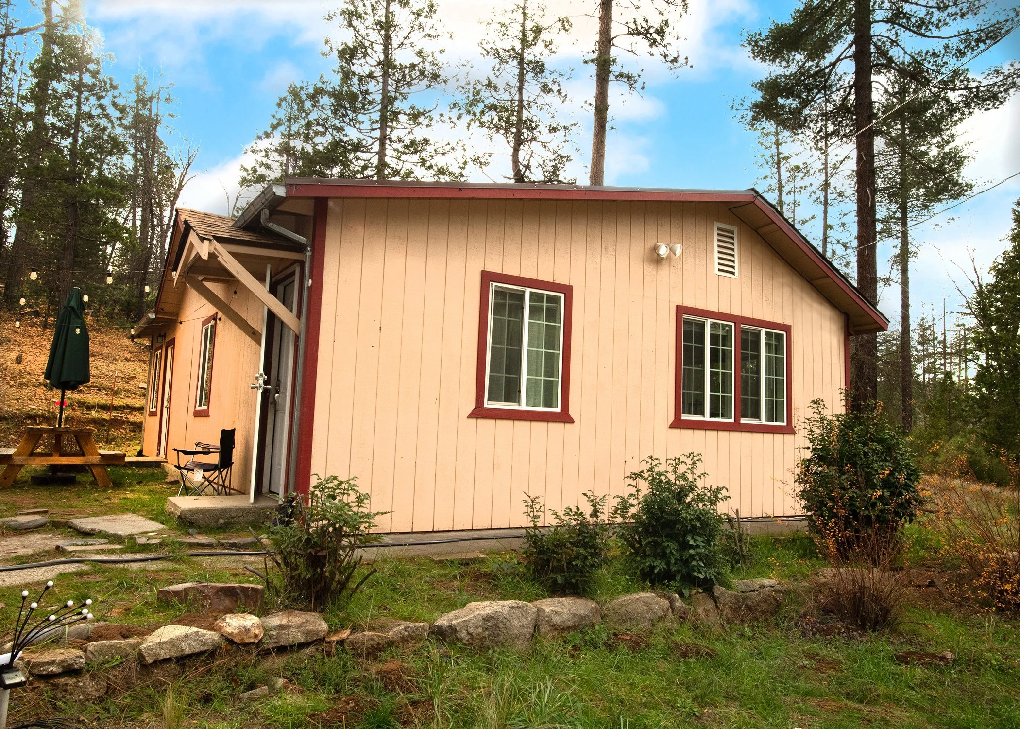 A small house painted beige with dark red trim, situated in a wooded area with tall trees, green grass, and bushes. The house has three windows, a small porch, a picnic table with an umbrella, and outdoor lighting.