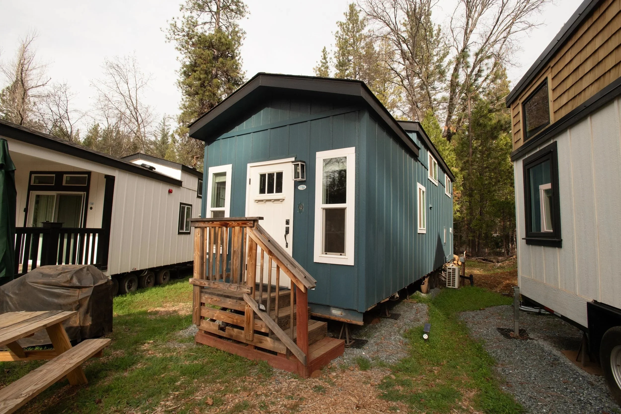 A small blue house with white window frames, a small set of wooden stairs leading to the front door, and surrounded by other houses or trailers in a wooded area.