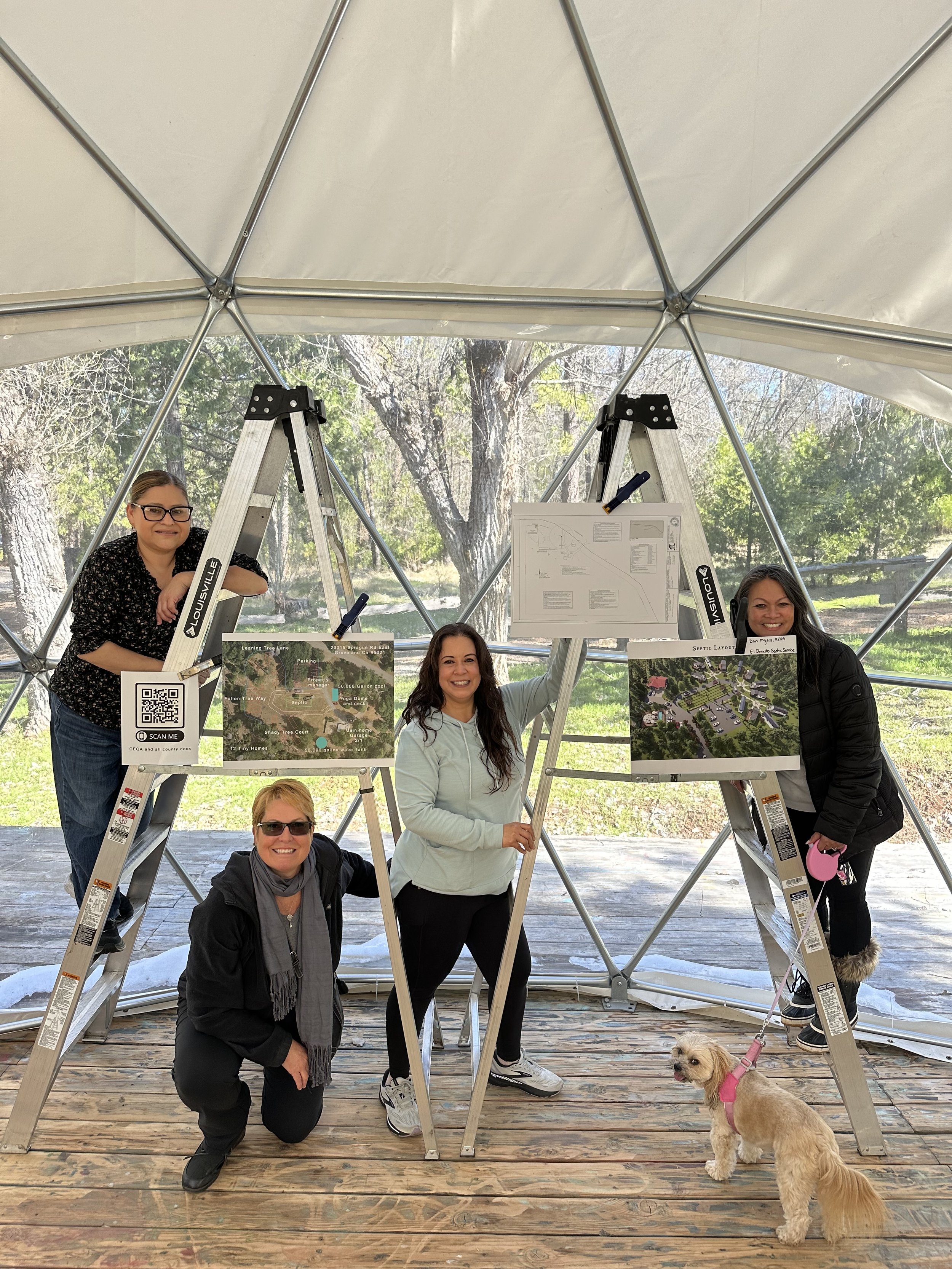Group of four women and a dog inside a geodesic dome with ladders, maps, and informational boards, outdoors with trees in the background.