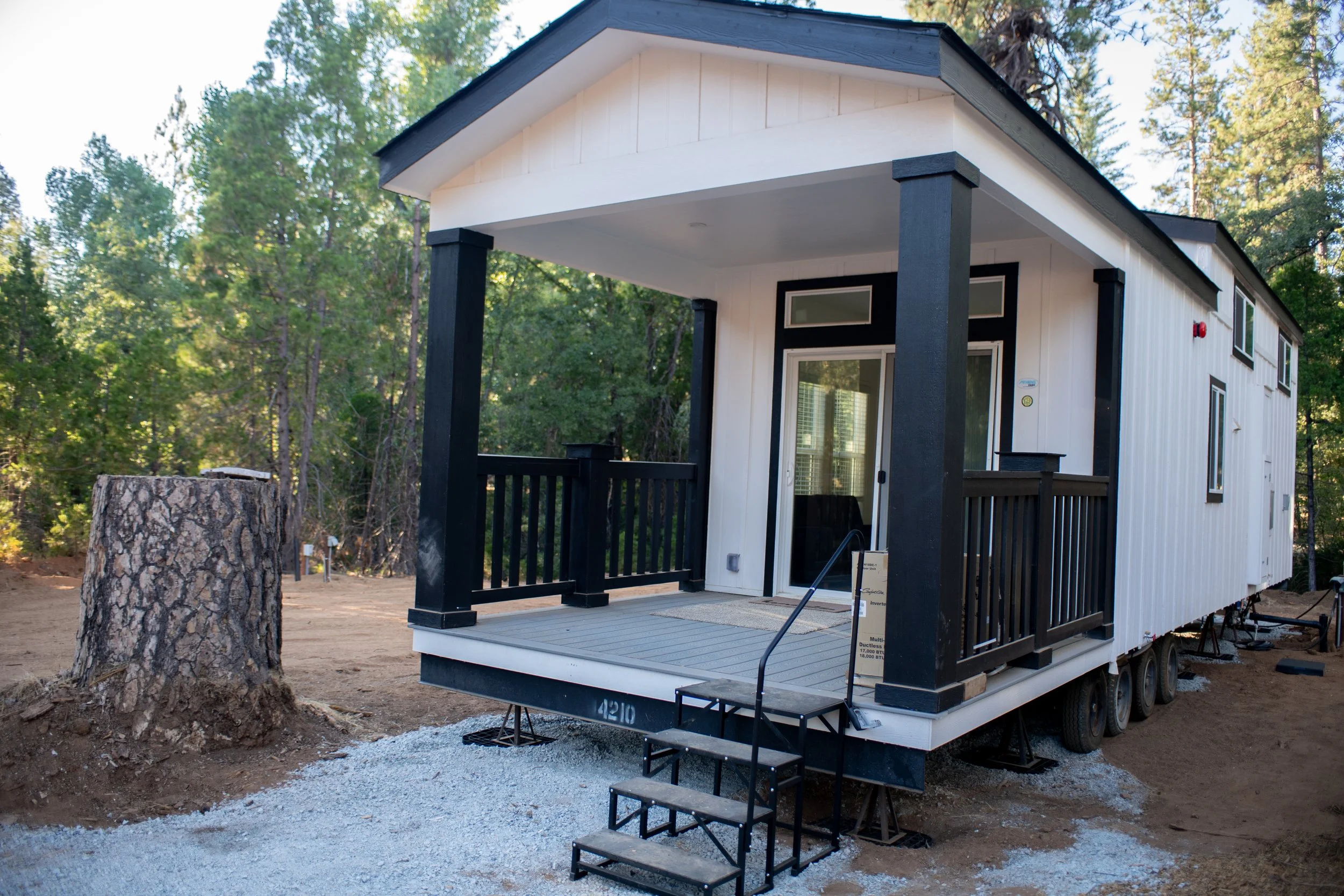 Newly constructed tiny house on wheels with white exterior and black accents, situated in a wooded area, with a small front porch and steps.