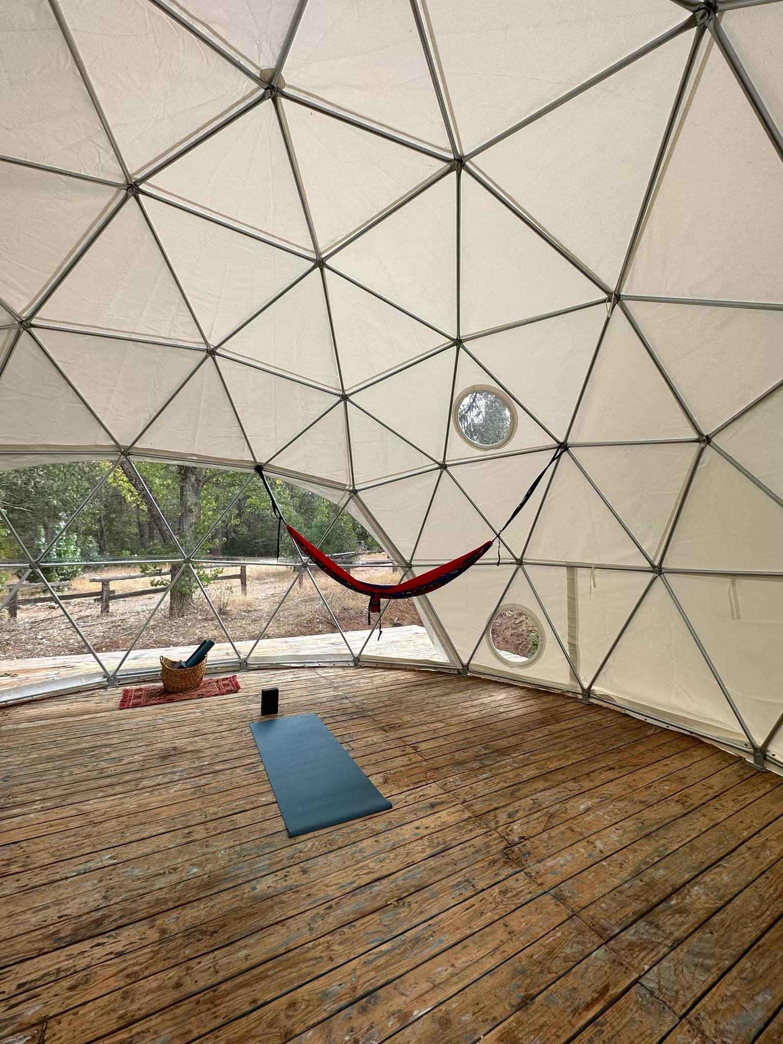 Interior of a geodesic dome with wooden floor, a yoga mat, a hammock, and small circular windows showing trees outside.