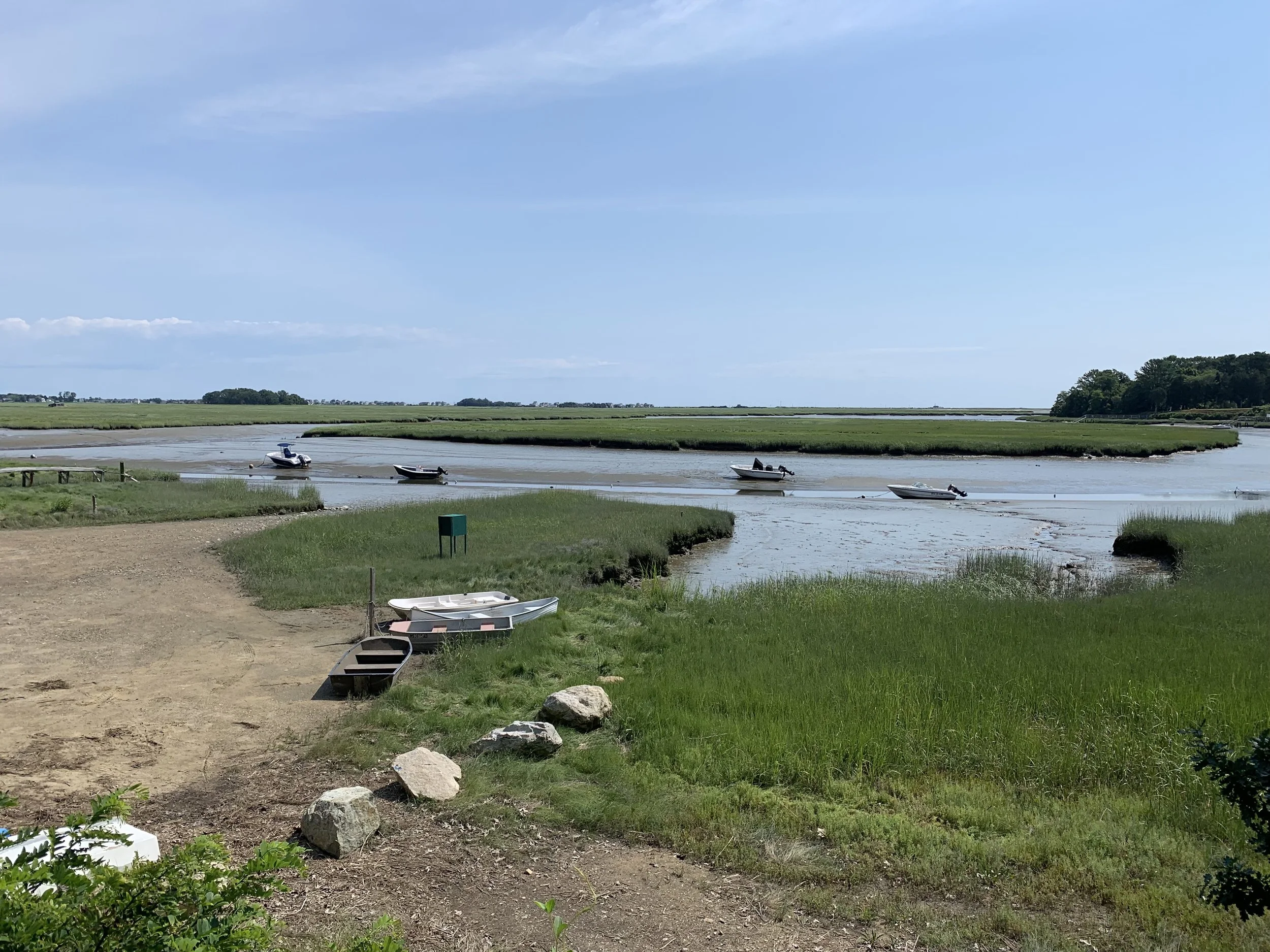 A beautiful estuary at low tide with dinghies, stretches of green marsh grass, mud flats, a small dock, and blue sky.