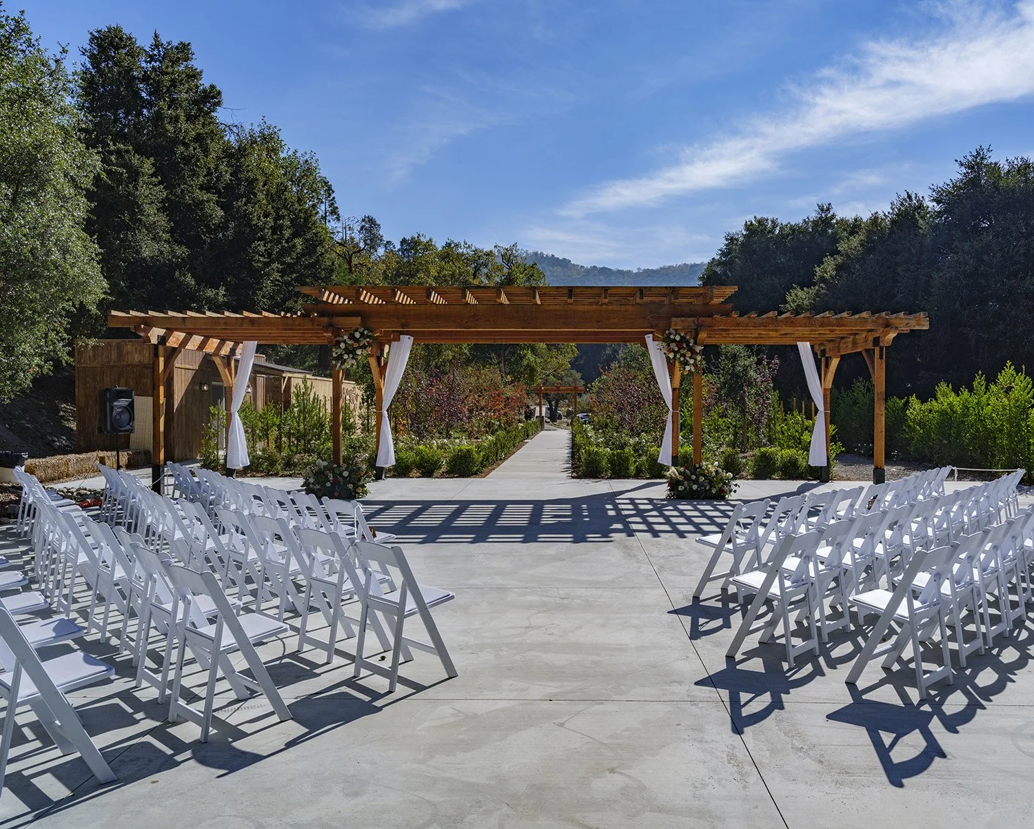 Outdoor wedding venue with white chairs arranged on a concrete patio, wooden archway decorated with flowers, surrounded by trees and mountains under a blue sky.
