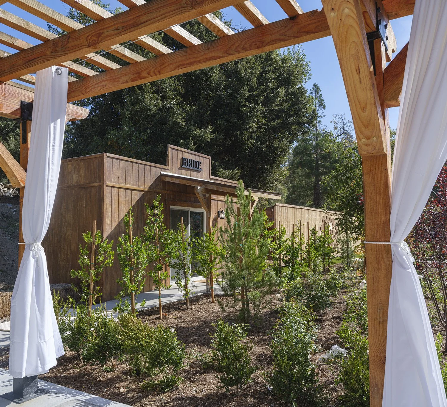 A wooden wedding arch with white curtains, overlooking a garden with small trees and a small wooden building labeled 'BRIDE' in the background.