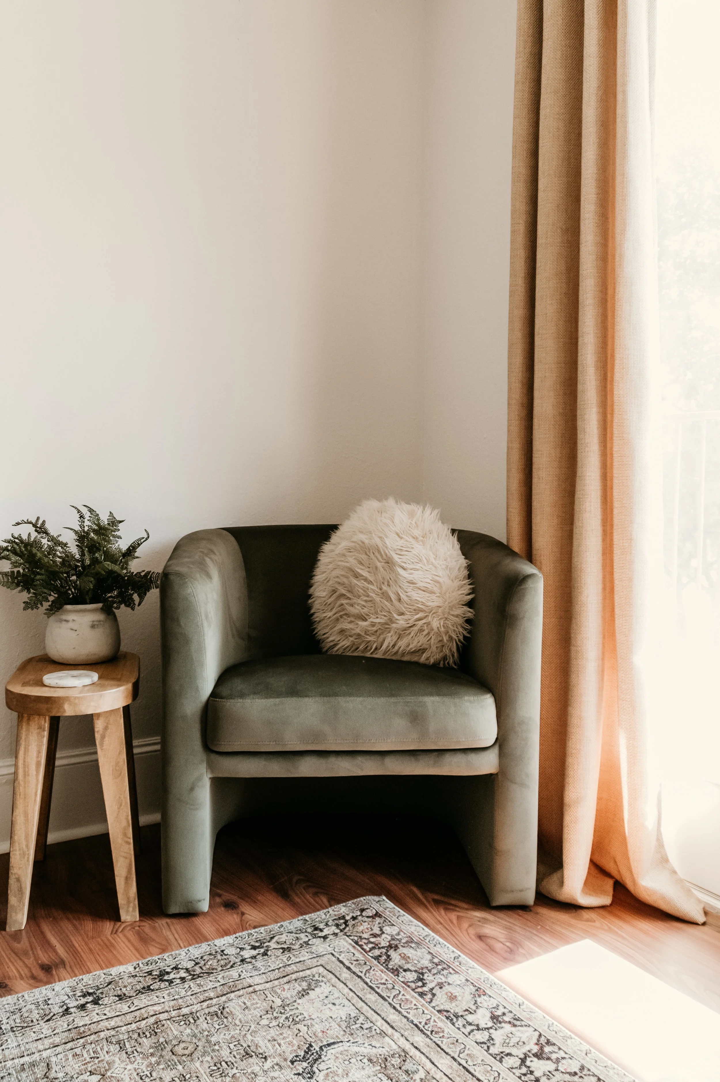 A cozy corner of a living room with a green velvet armchair, a fluffy beige pillow, a wooden side table with a potted plant, a patterned area rug on a hardwood floor, cream-colored walls, and peach curtains near a window.