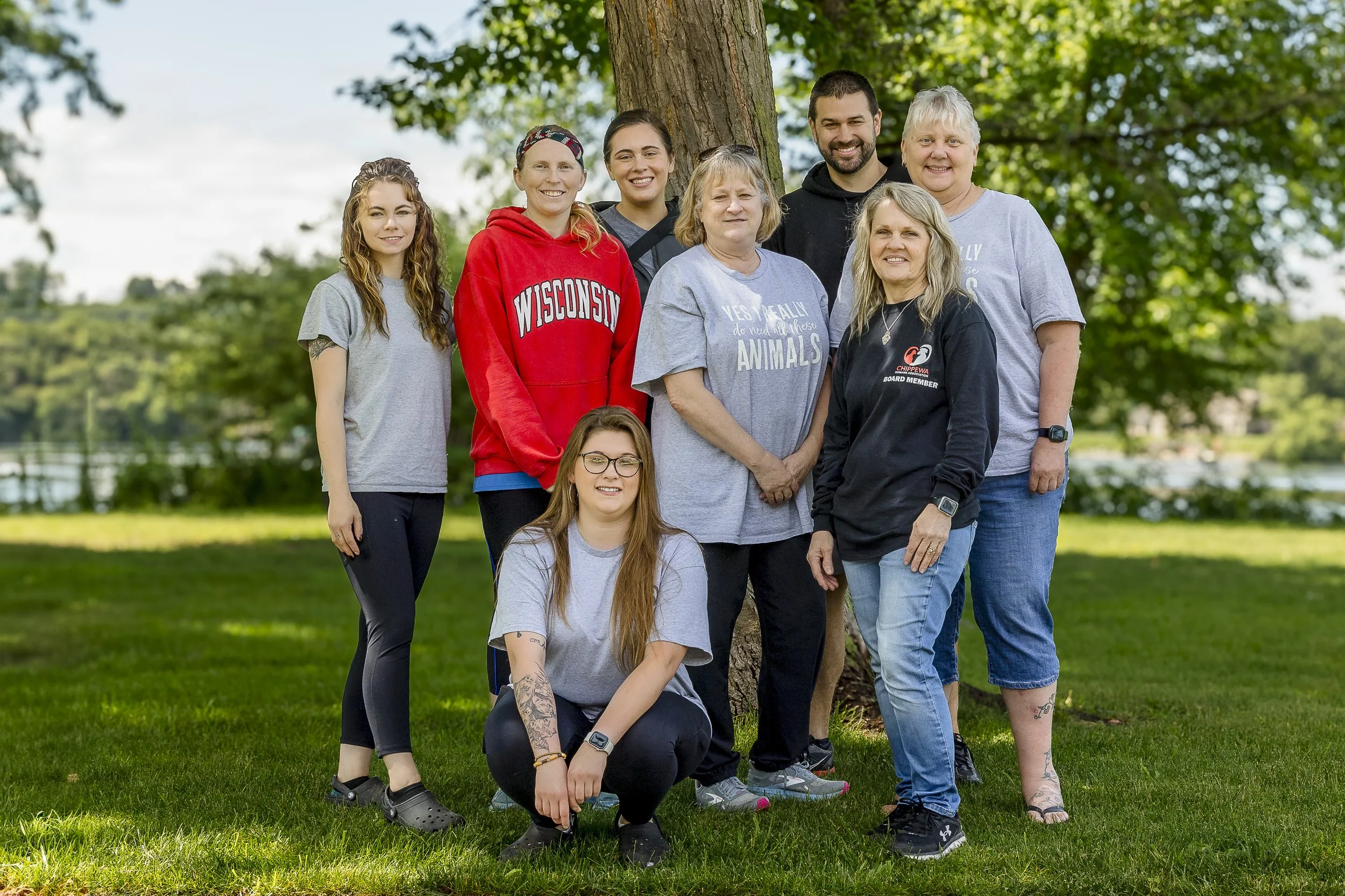 Group of nine diverse women and girls standing outdoors by a tree, smiling, with a lake and trees in the background.