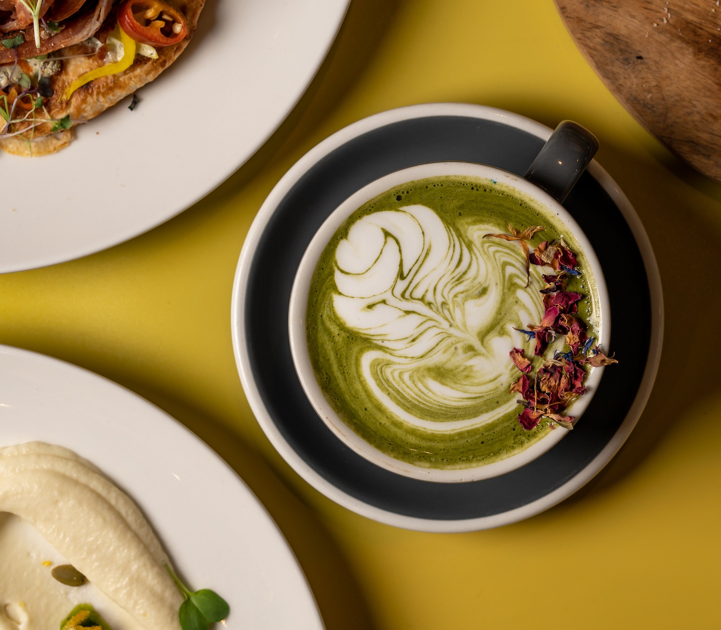 Matcha latte art with rose petals in a cup next to plated dishes on a yellow table.