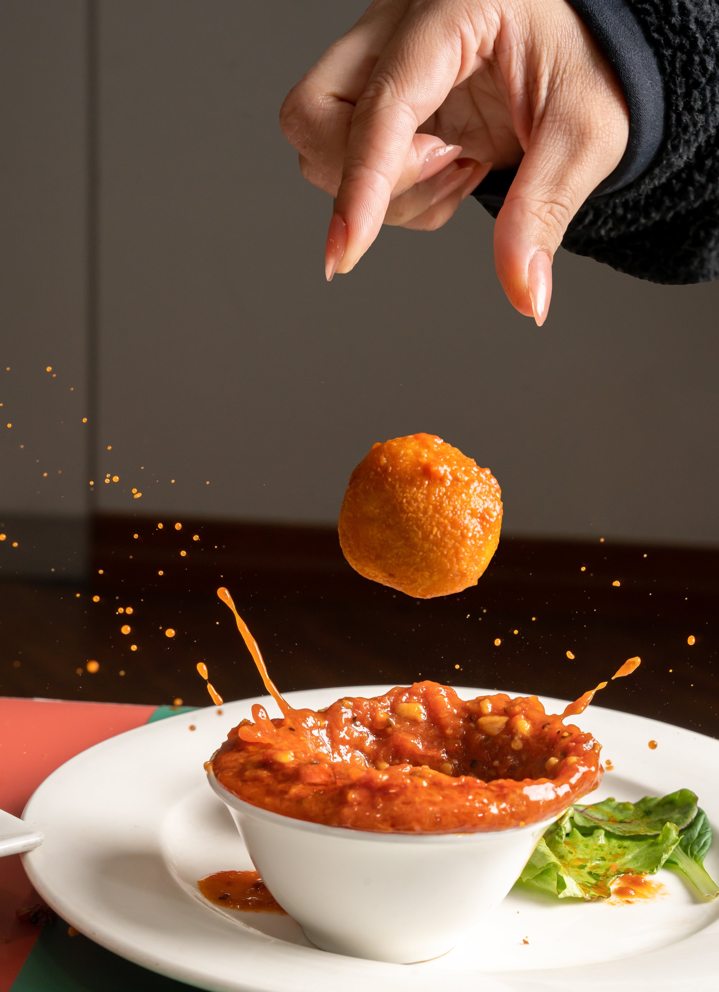 Hand dropping a fried ball into a bowl of spicy sauce, causing a splash, on a white plate with lettuce.