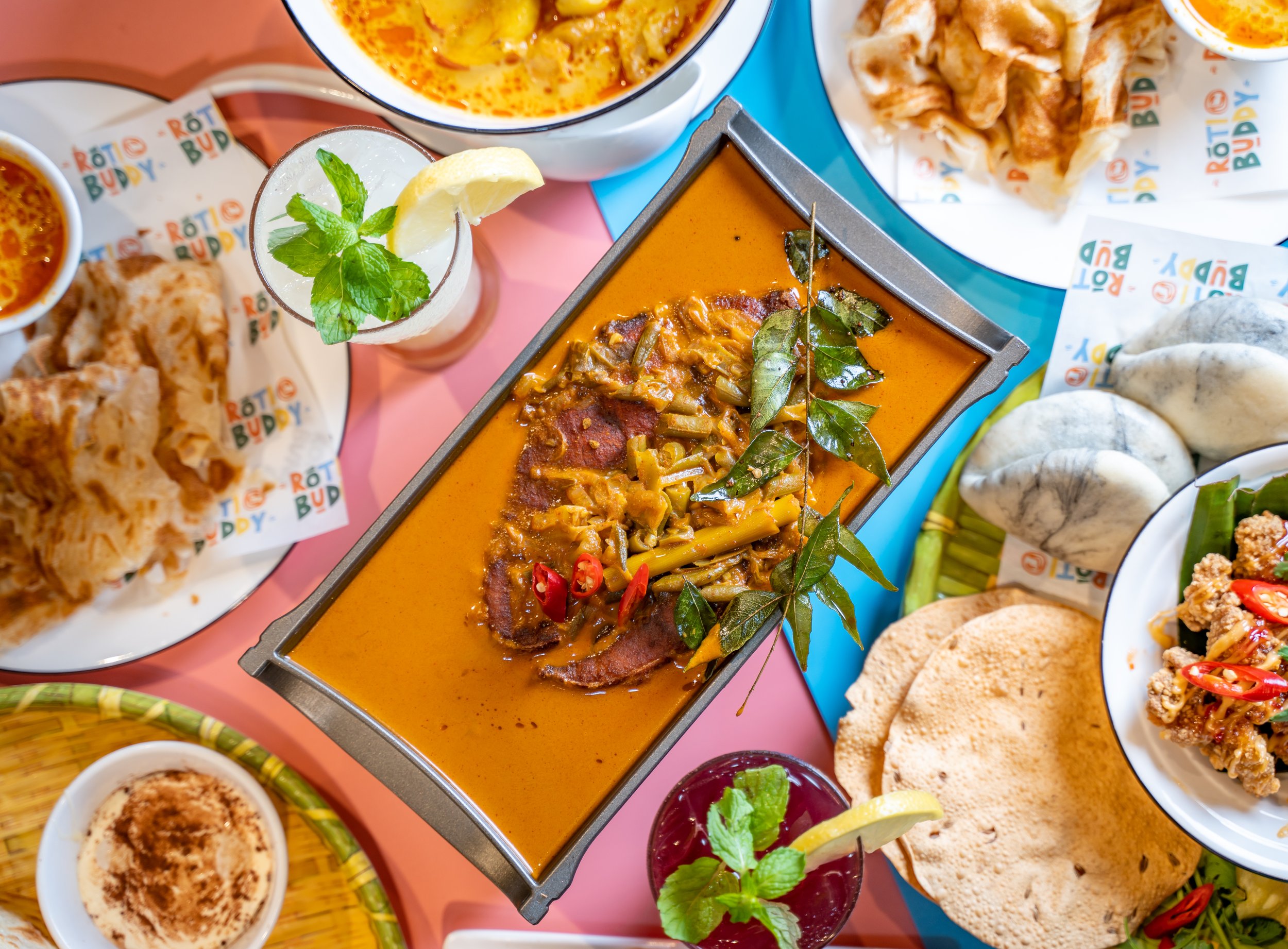 Top view of assorted Asian dishes, including curry, crispy bread, and beverages with herbs on a colorful table.