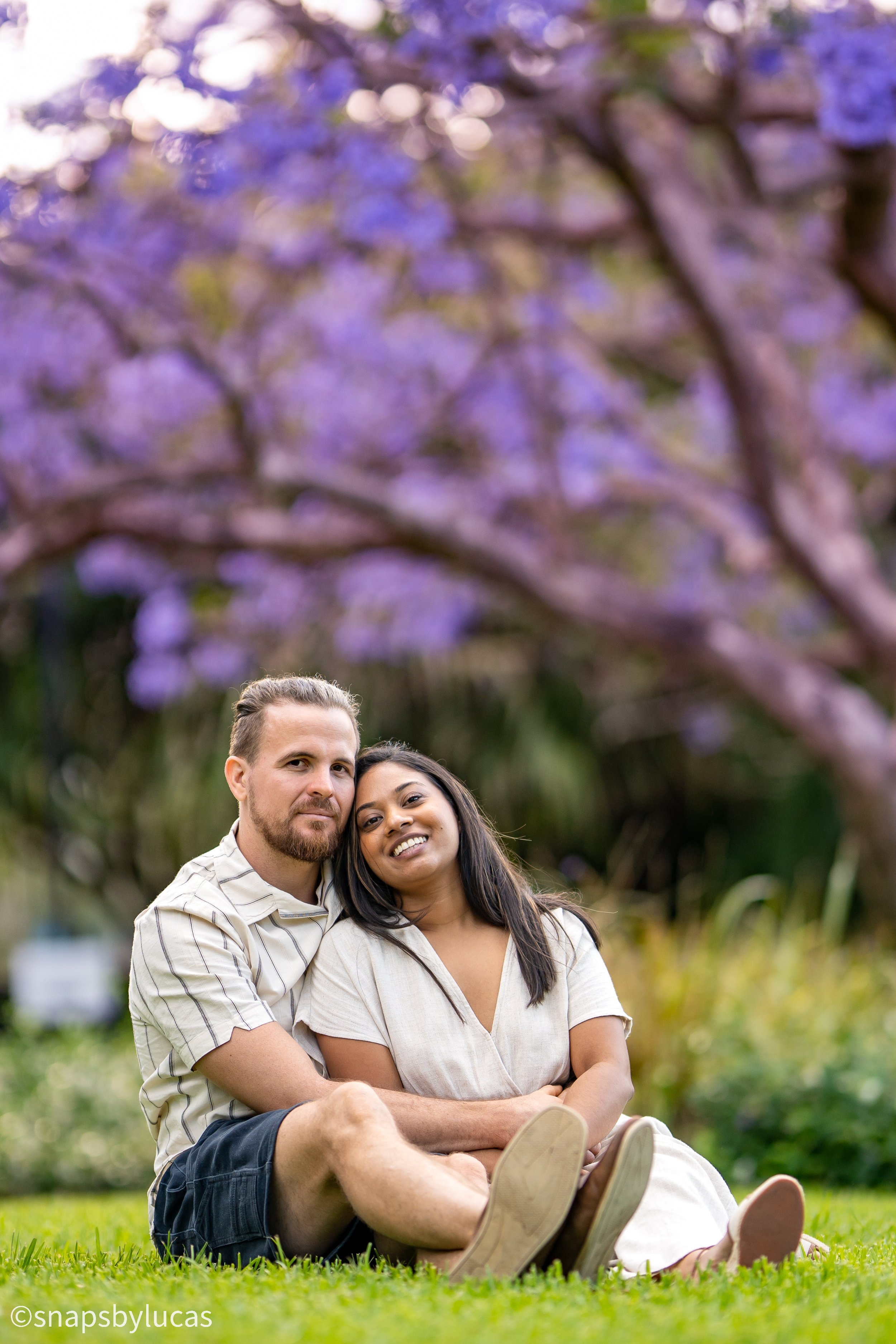 Couple sitting on grass under purple flowering tree