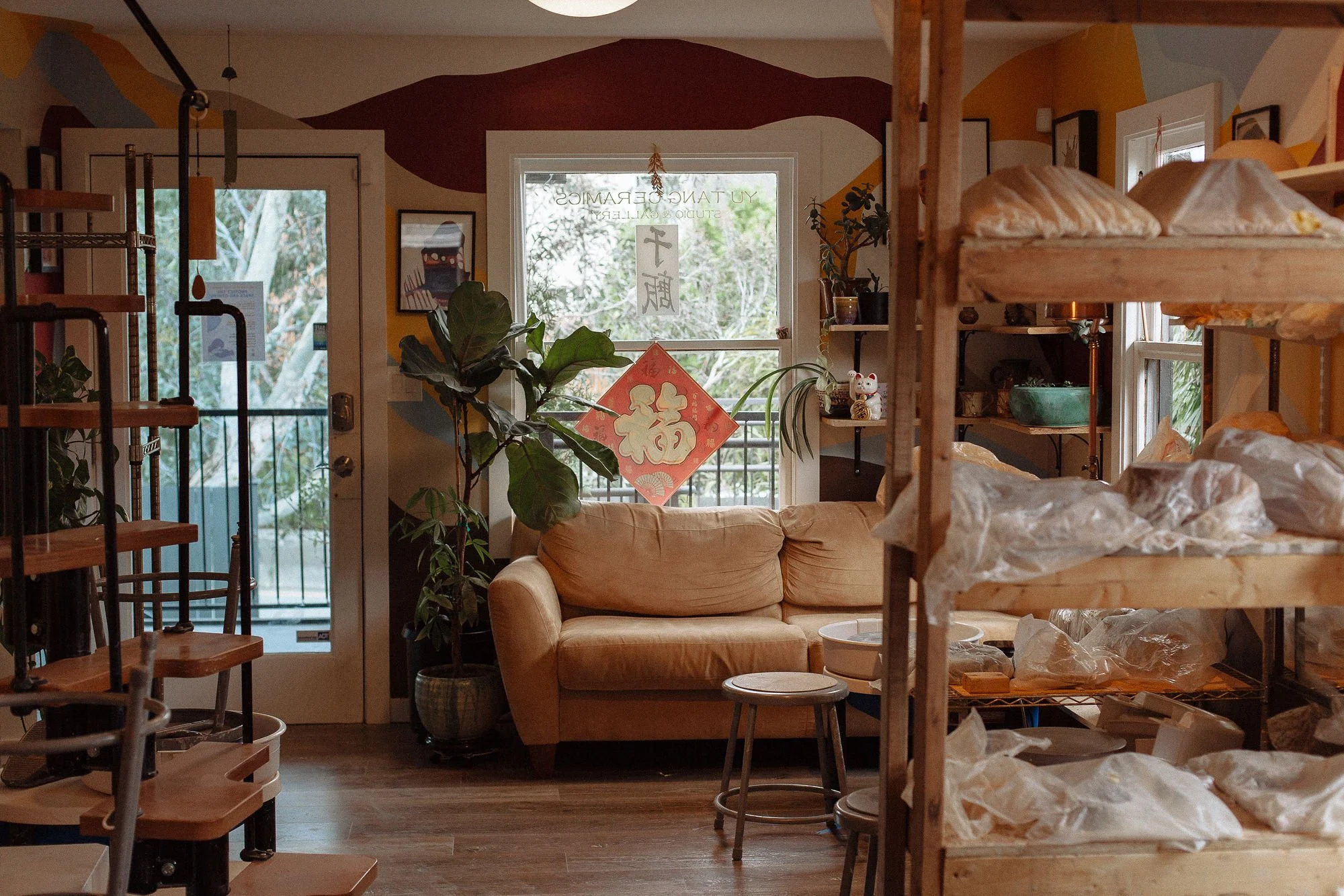 Interior of a cozy, artsy cafe or shop with shelves, a beige couch, potted plants, a large window with decorative signs, and wooden furniture.