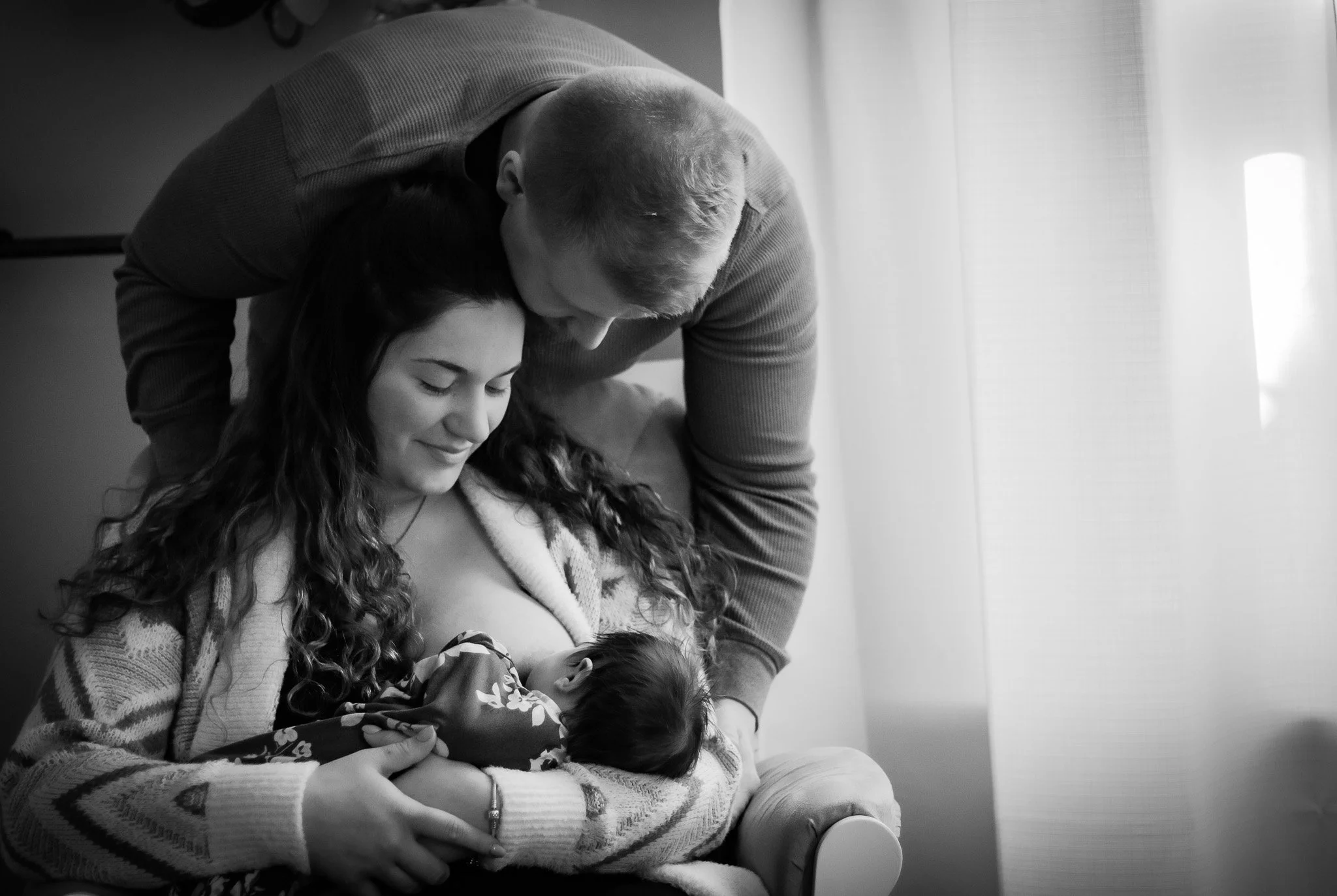 A mother and father look down lovingly at their newborn baby while she breastfeeds.