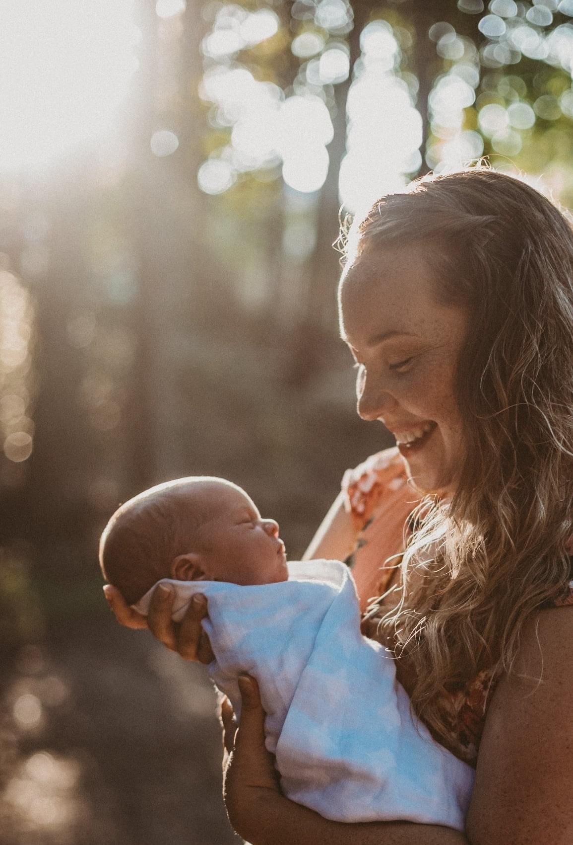 A mother and doula holds her newborn baby while standing in the woods. She smiles down at the baby wrapped in a light blue blanket, while sunlight shines on both of them.