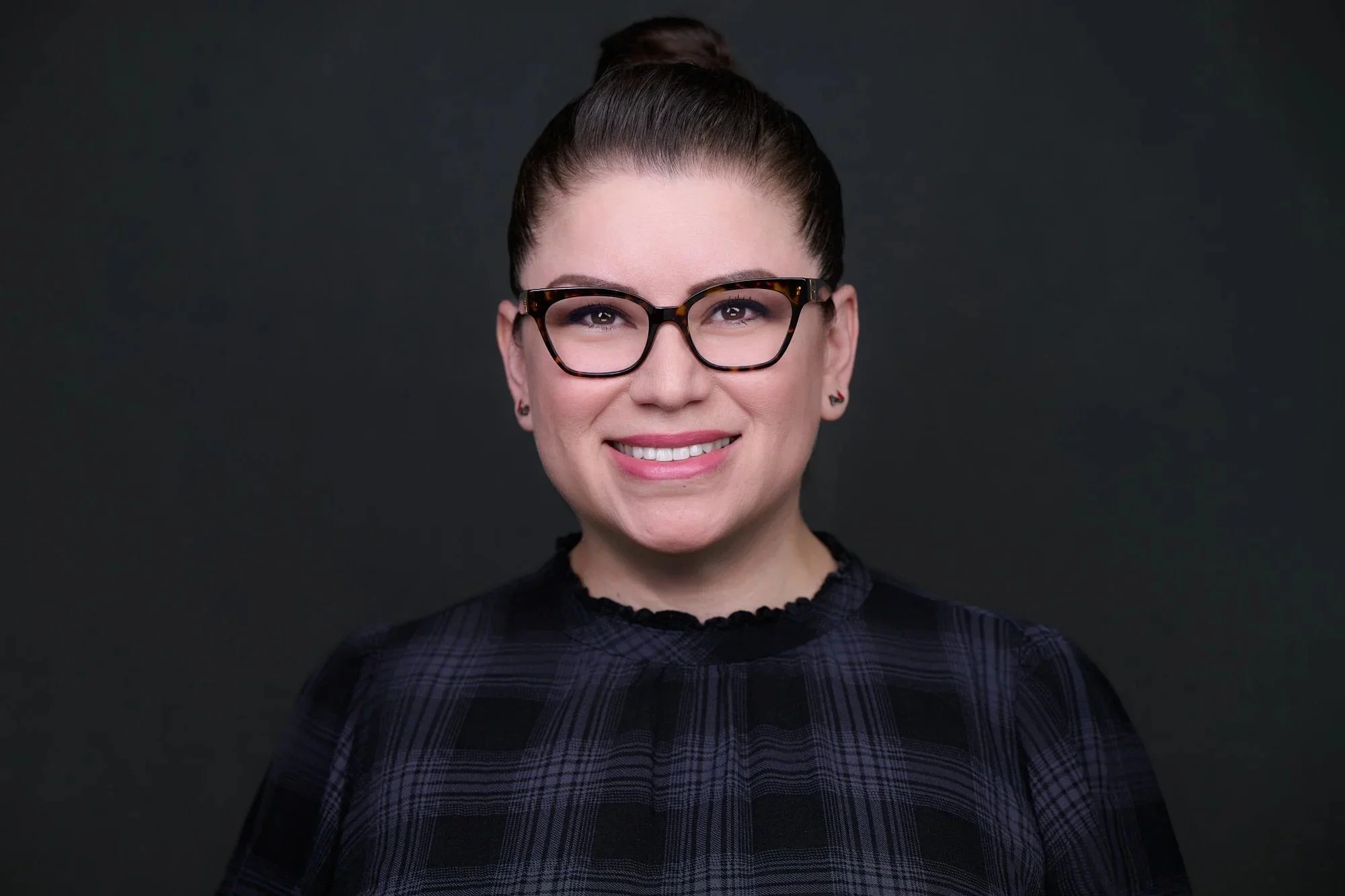 Corporate headshot of a smiling woman with glasses in a dark plaid top on a charcoal background.