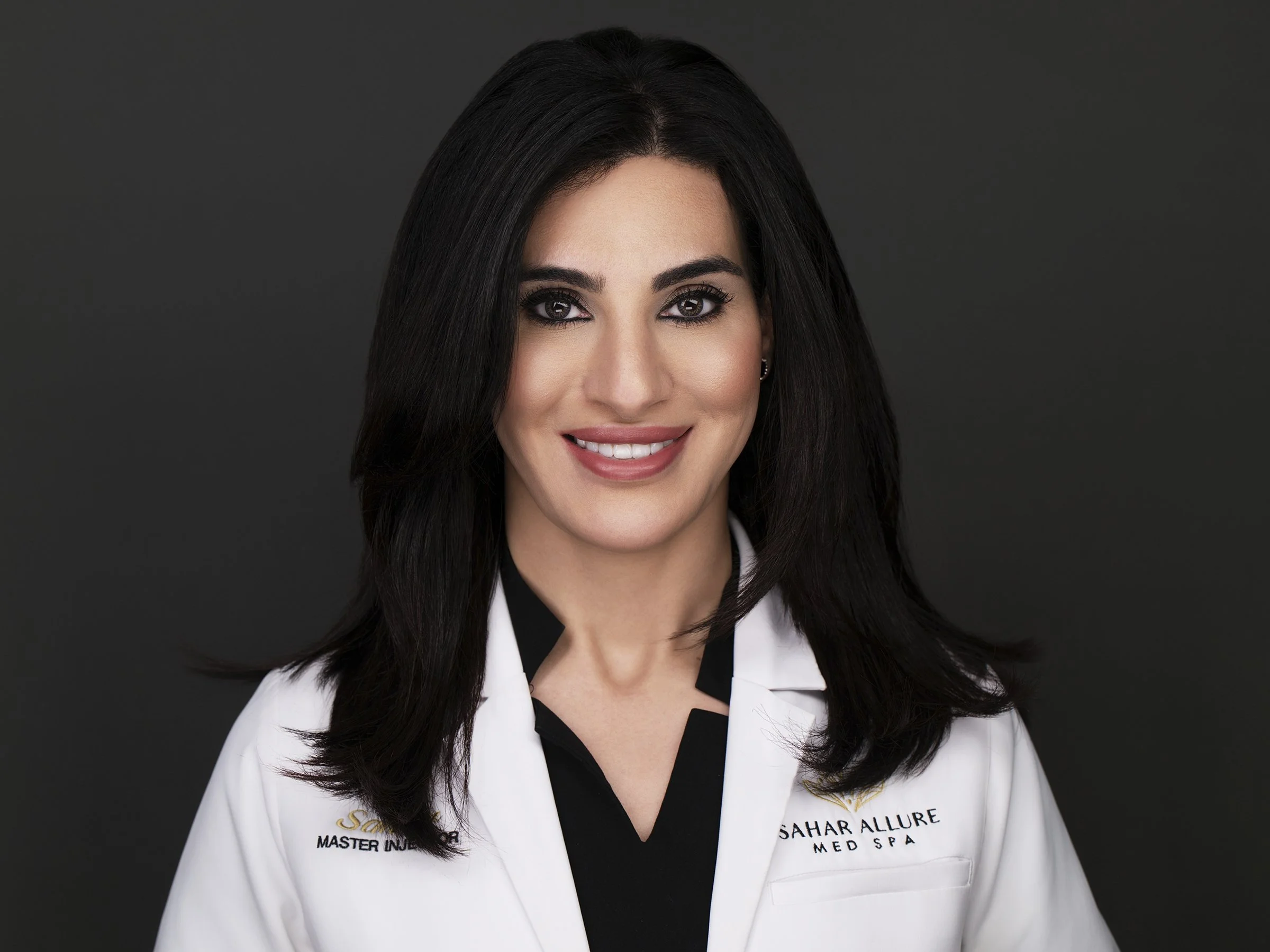 Medical headshot of a master injector with long dark hair in an embroidered white coat, photographed on a dark background.