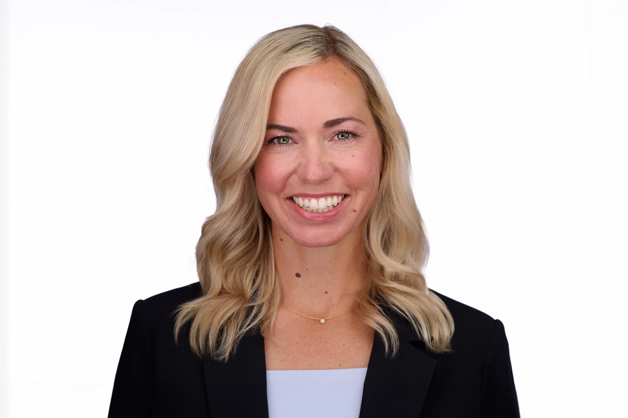 Business headshot of a woman with wavy blonde hair in a black blazer, photographed on a white background.