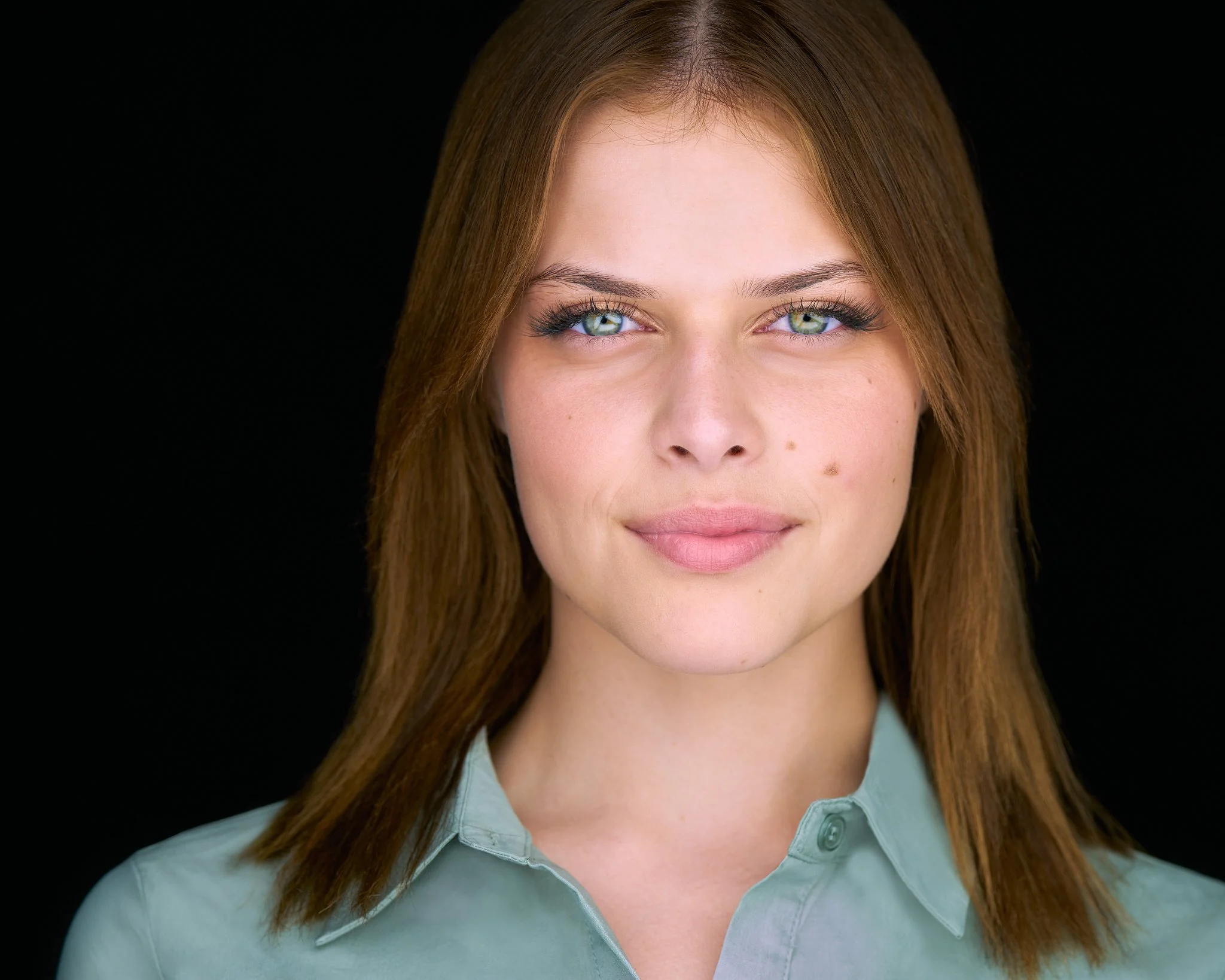 Headshot of a woman with long auburn hair in a sage-green shirt on a black background.