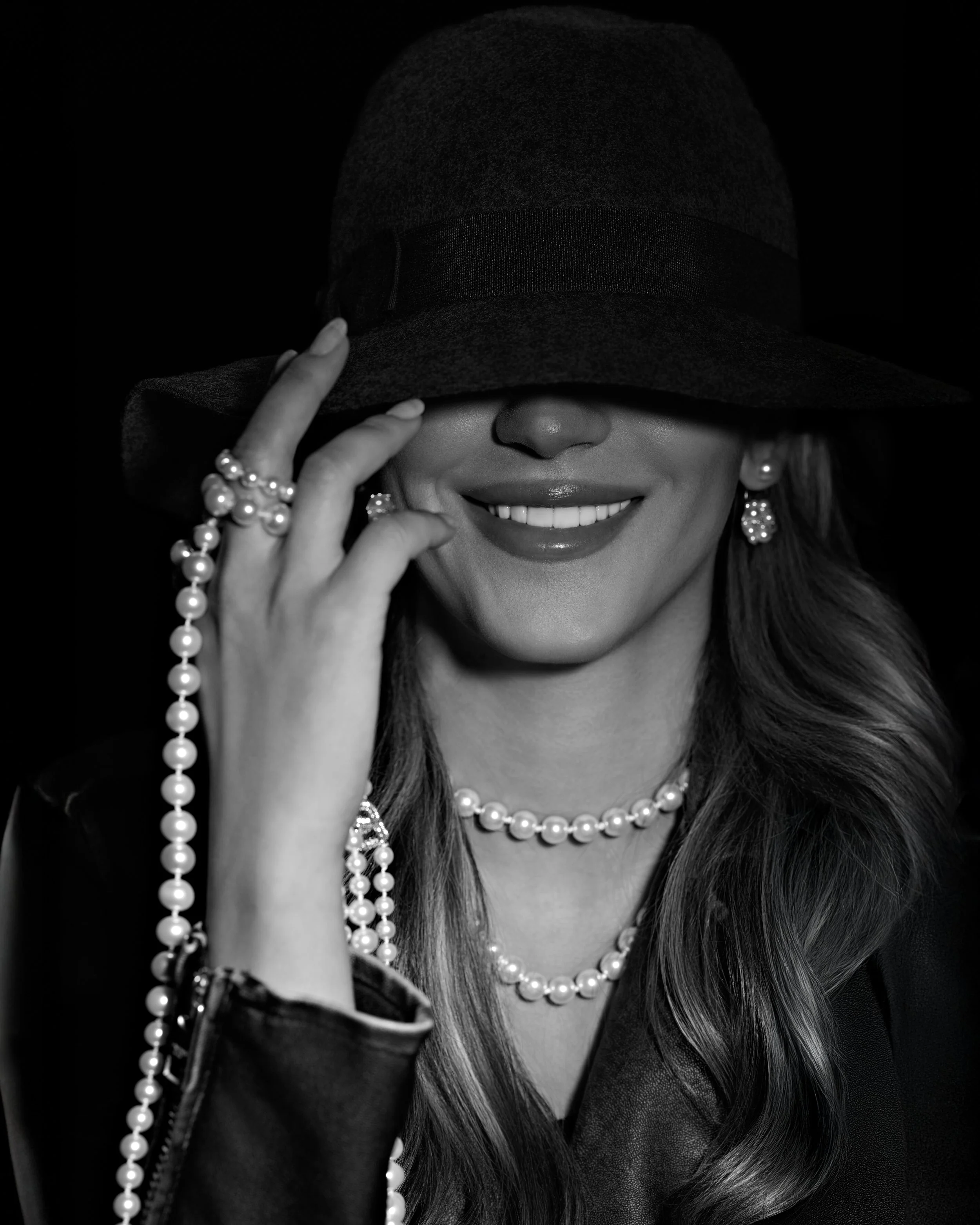 Black-and-white portrait of a smiling woman in a black hat and layered pearls