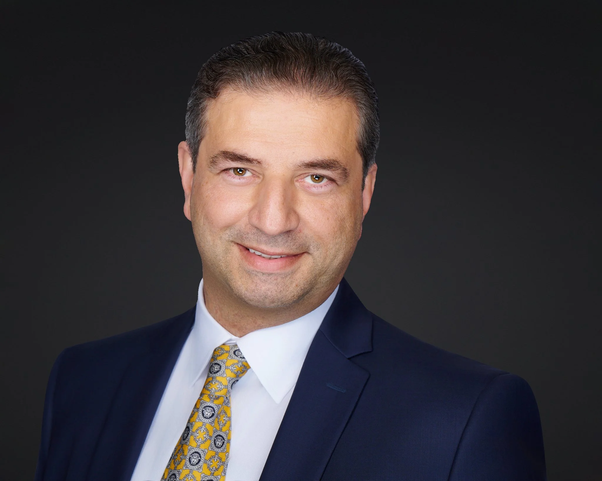 Attorney headshot of a man in a navy suit and gold tie on a dark background.