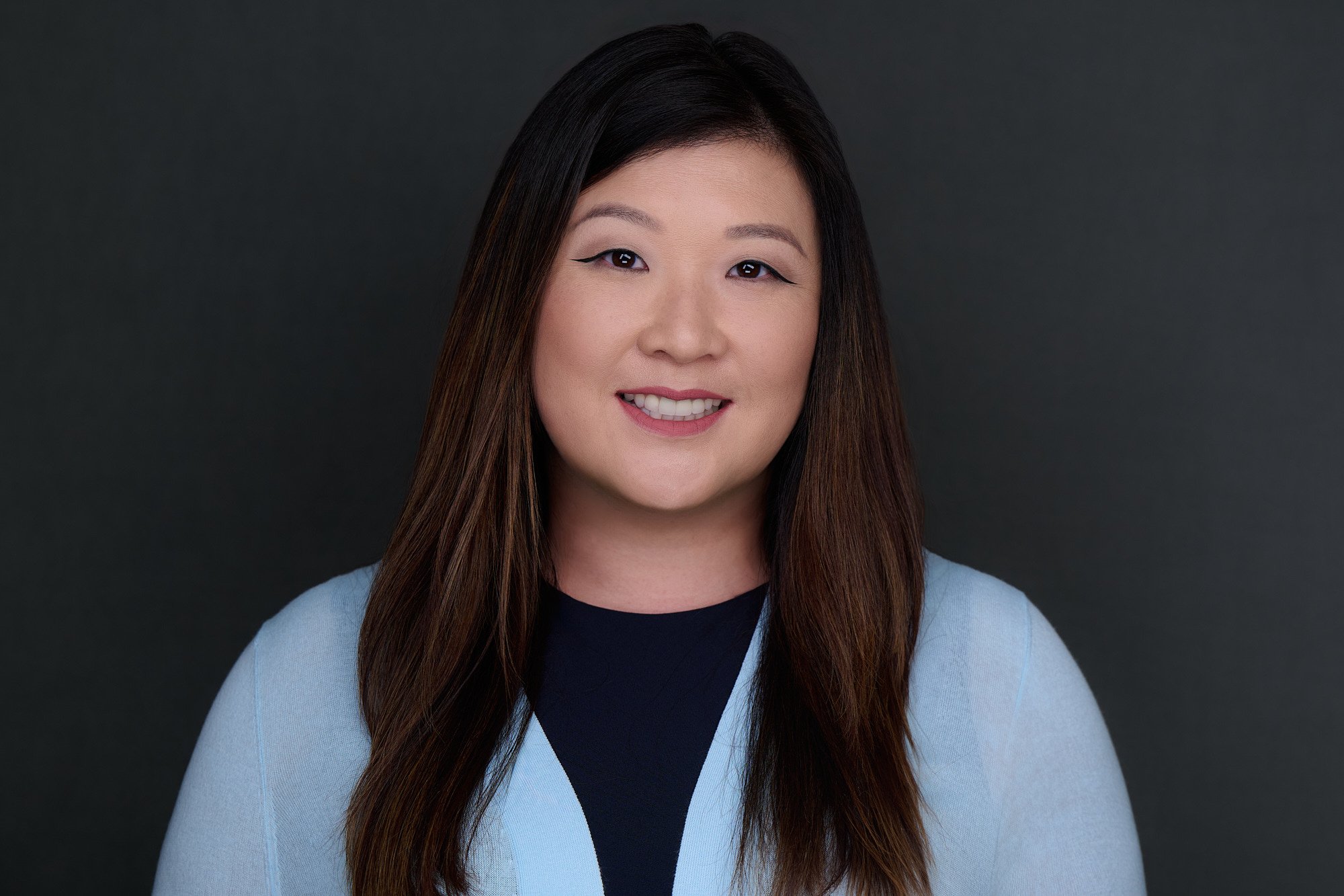 Corporate headshot of a woman in a light blue blazer on a charcoal backdrop, photographed on location in Orange County