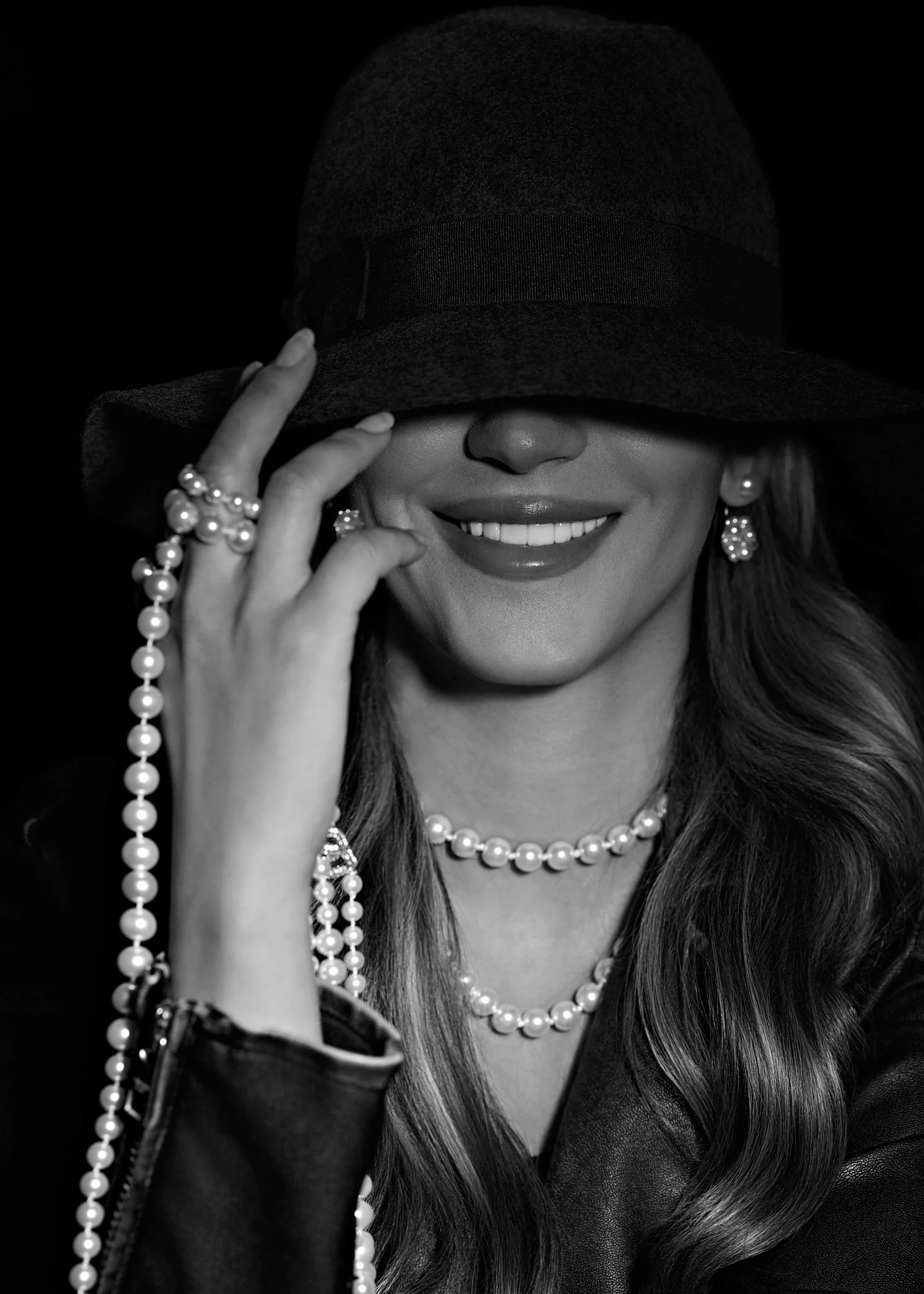 Black and white studio portrait of a laughing woman in a wide-brimmed hat and layered pearls.