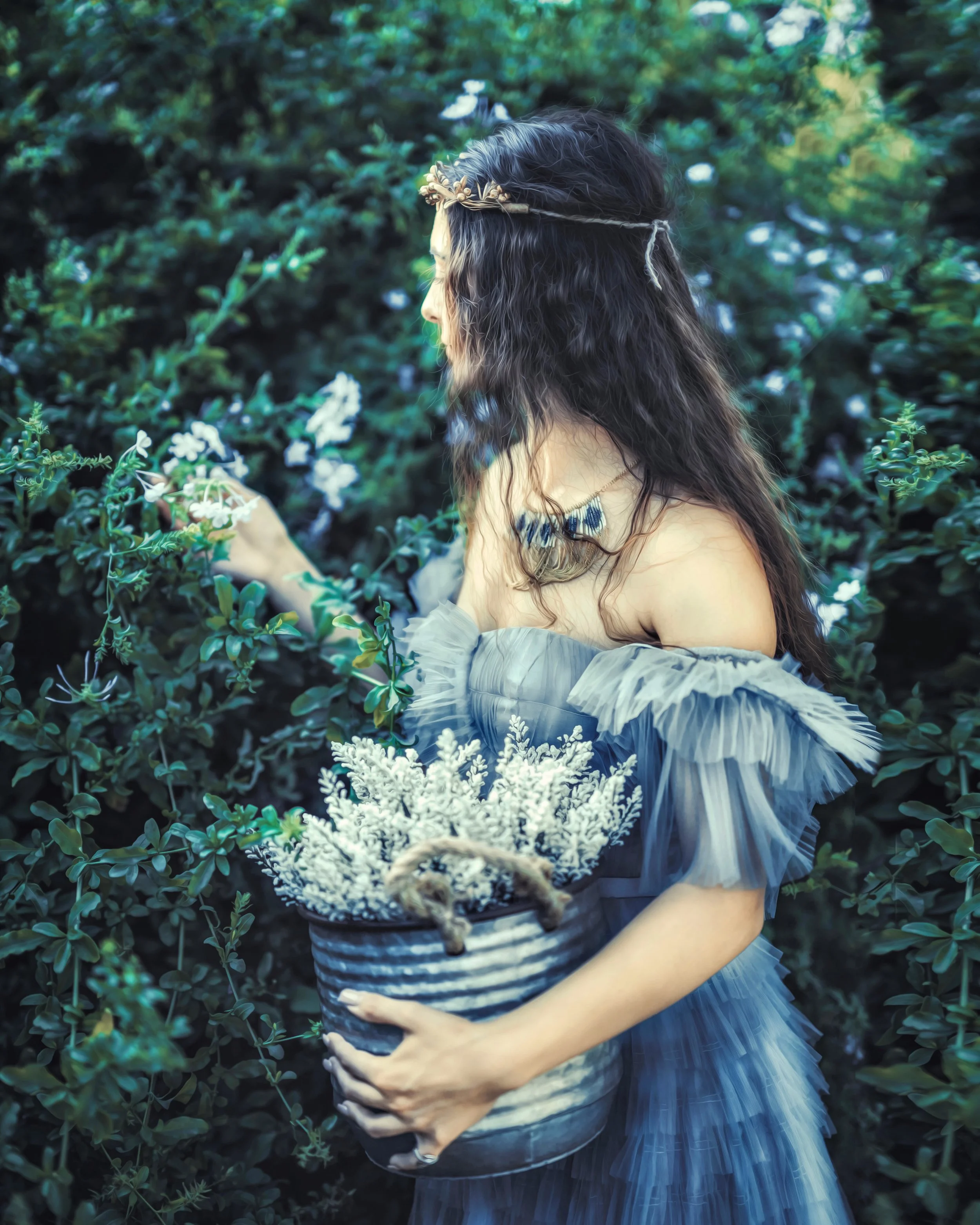 Outdoor portrait of a woman in a blue tulle dress and gold headpiece, holding a bucket of white flowers in a garden.