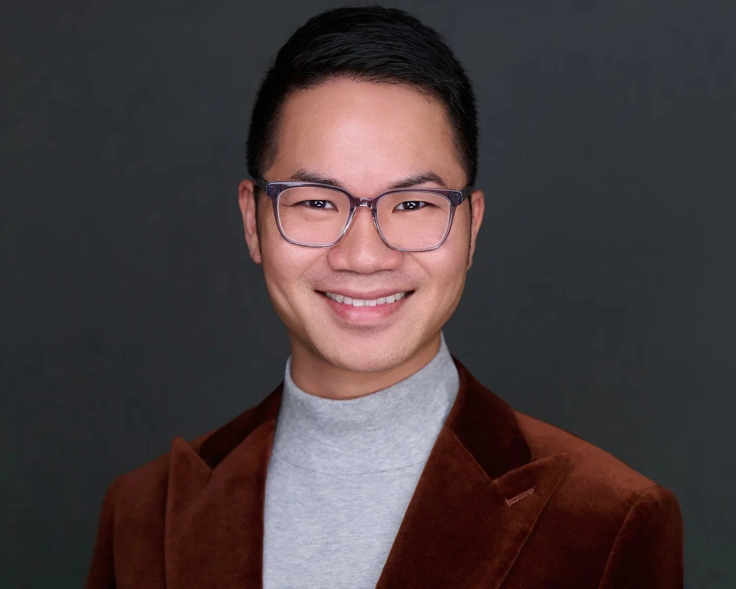 Corporate headshot of a smiling man in glasses and a rust velvet blazer on a charcoal background.