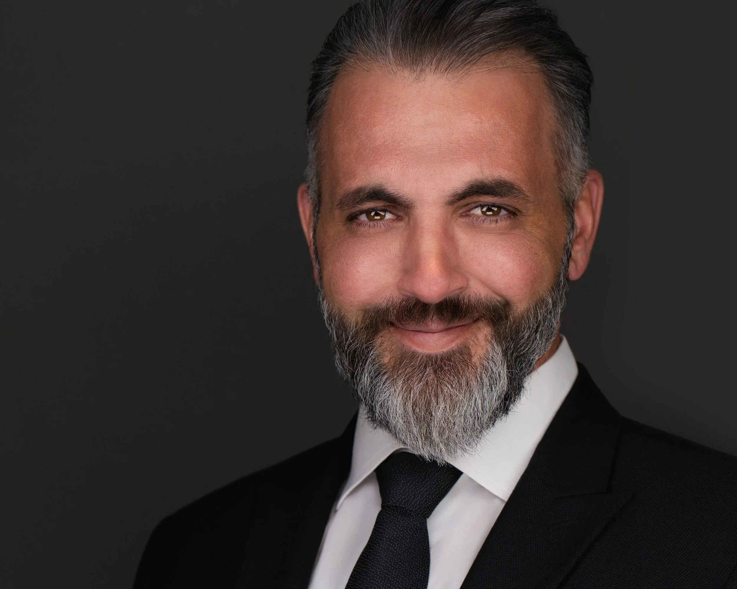 Executive headshot of a man with a grey beard in a black suit, white shirt and black tie, photographed on a dark grey background.