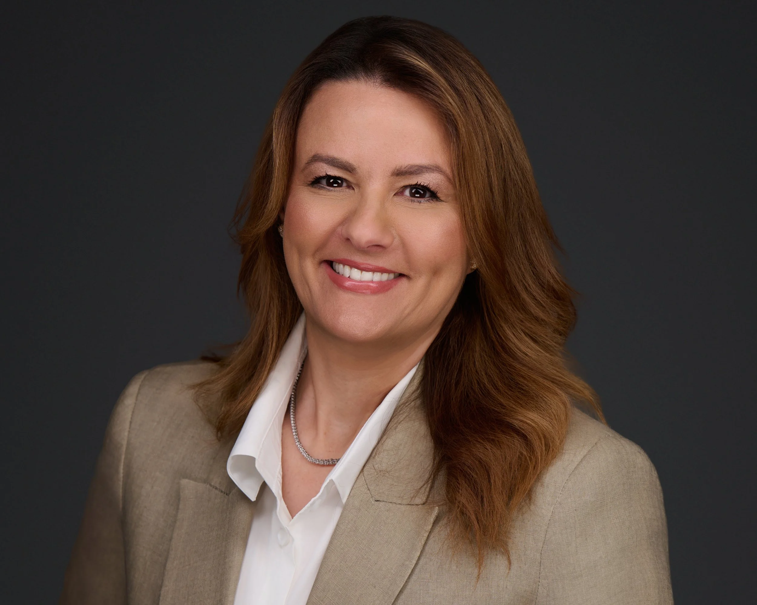 Studio headshot of a woman in a taupe blazer and white shirt against a dark grey background.