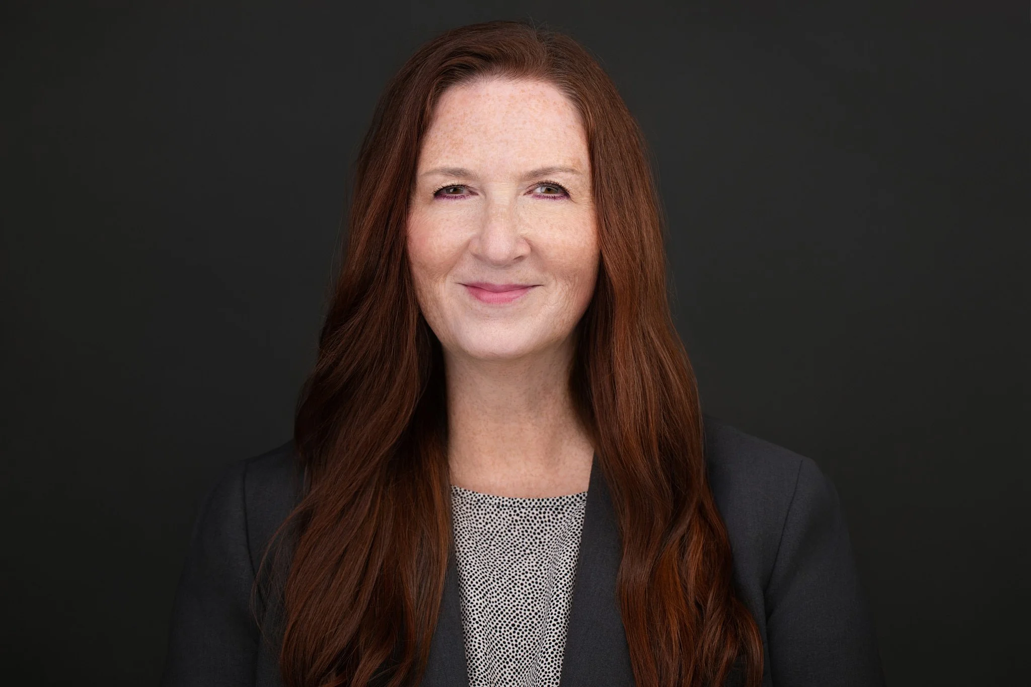 Business headshot of a woman with long auburn hair in a grey top and dark blazer.