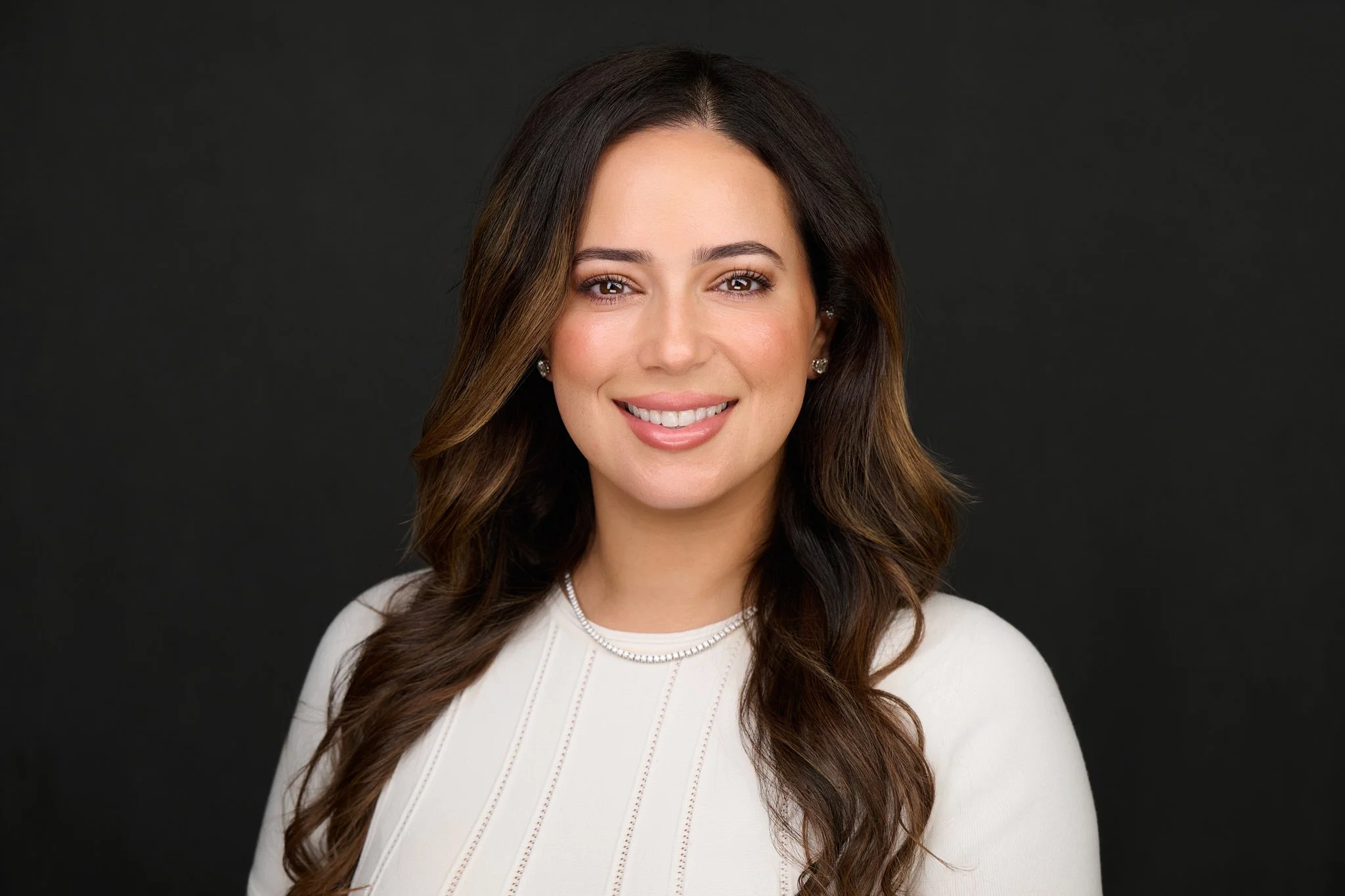 Business headshot of a woman with long dark wavy hair in a cream knit top, photographed on a dark background.